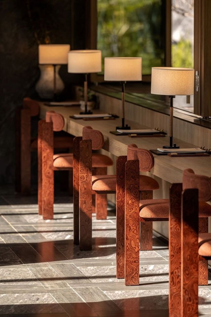 A row of wooden chairs with upholstered seats beside a long desk with table lamps and open notebooks, near large windows with sunlight streaming in.