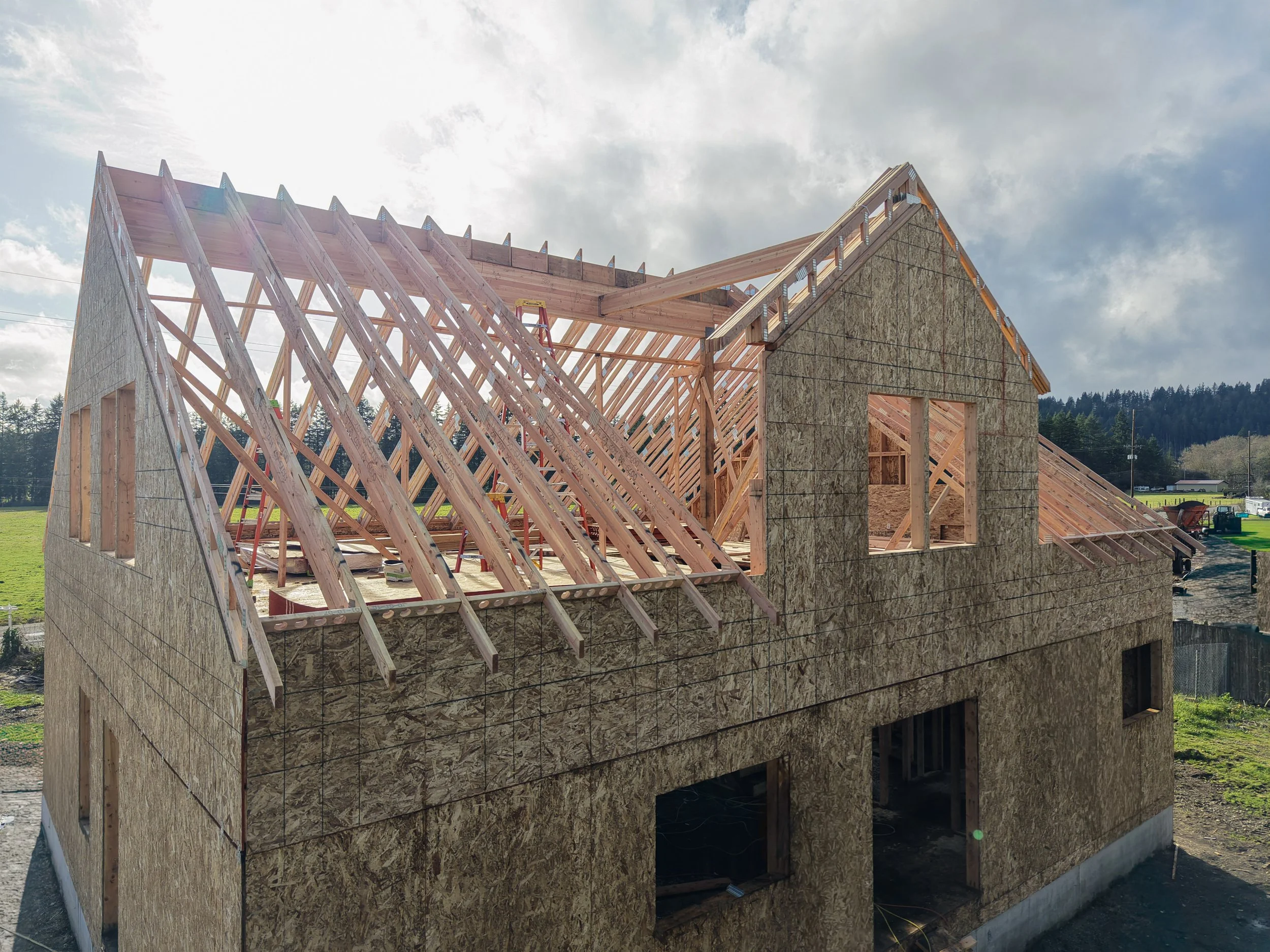 Construction site with a house framing in progress, showing wooden roof trusses and wall sheathing, under a partly cloudy sky.