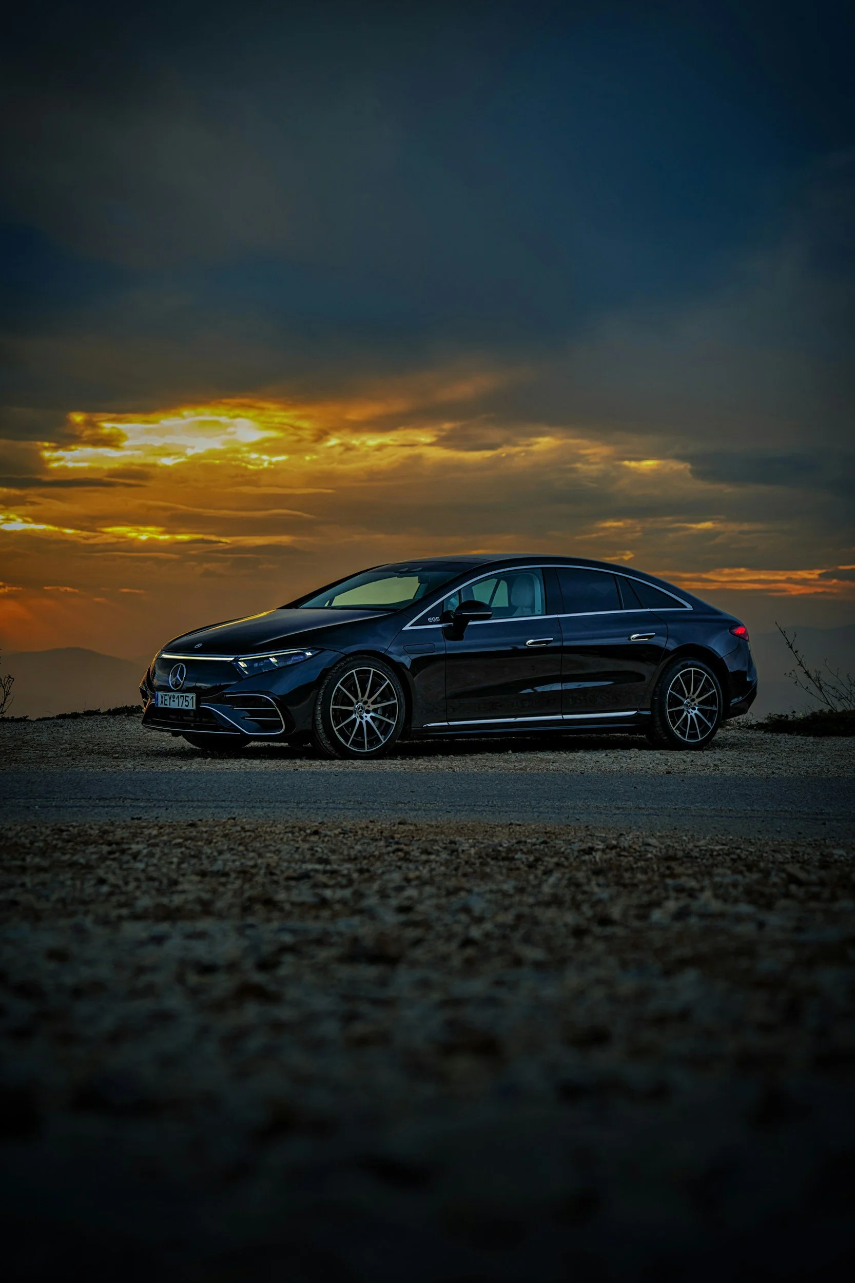 A black Mercedes-Benz sedan parked on a gravel surface at sunset with a dramatic sky filled with clouds in the background.