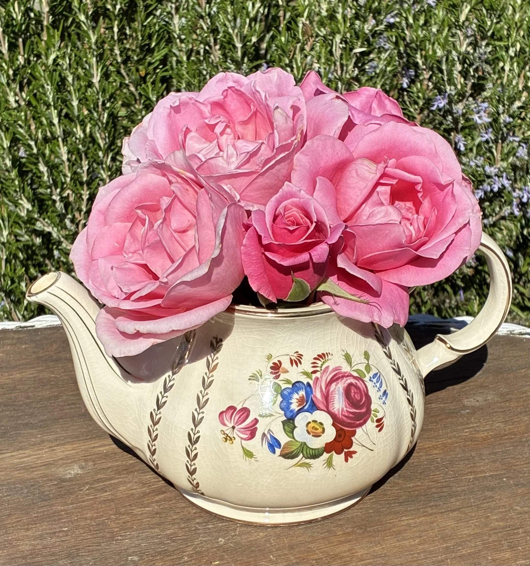 A vintage teapot with floral design, filled with pink roses, placed on a wooden surface outdoors with green foliage in the background.