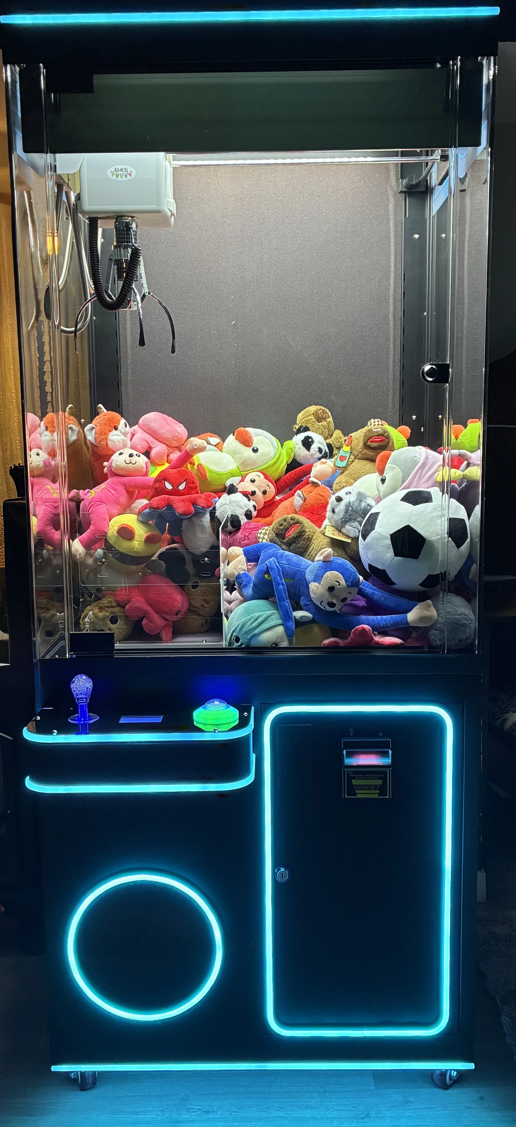 Claw machine filled with plush toys, including soccer balls, animals, and cartoon characters, with blue neon accents.