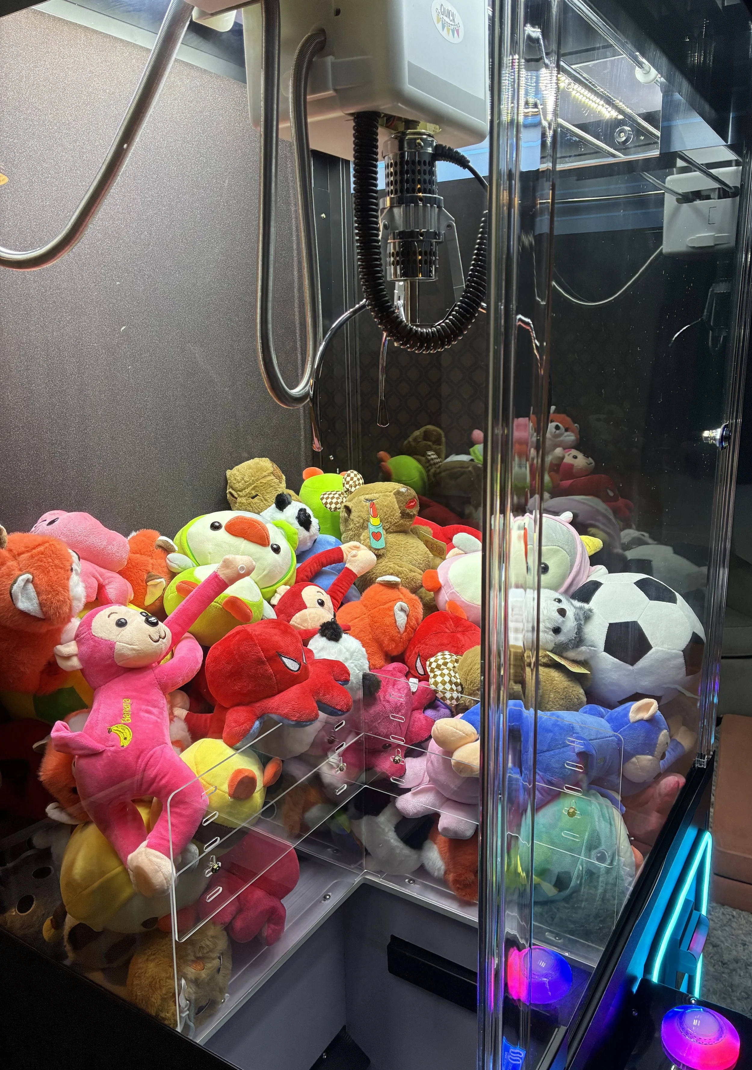 Claw machine filled with colorful plush toys, including a pink monkey, red octopus, green frog, soccer ball, and various other stuffed animals.