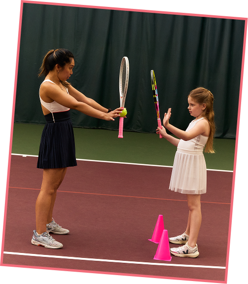 An adult woman and a young girl are practicing tennis on an indoor court. The woman is holding a tennis racket and ball, while the girl holds a racket with an instructor stick. They are standing behind pink cones.