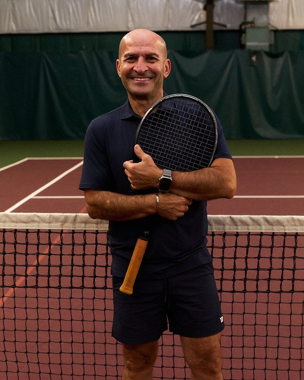 A man standing on an indoor tennis court, smiling and holding a tennis racket, wearing a dark polo shirt, shorts, and a smartwatch.