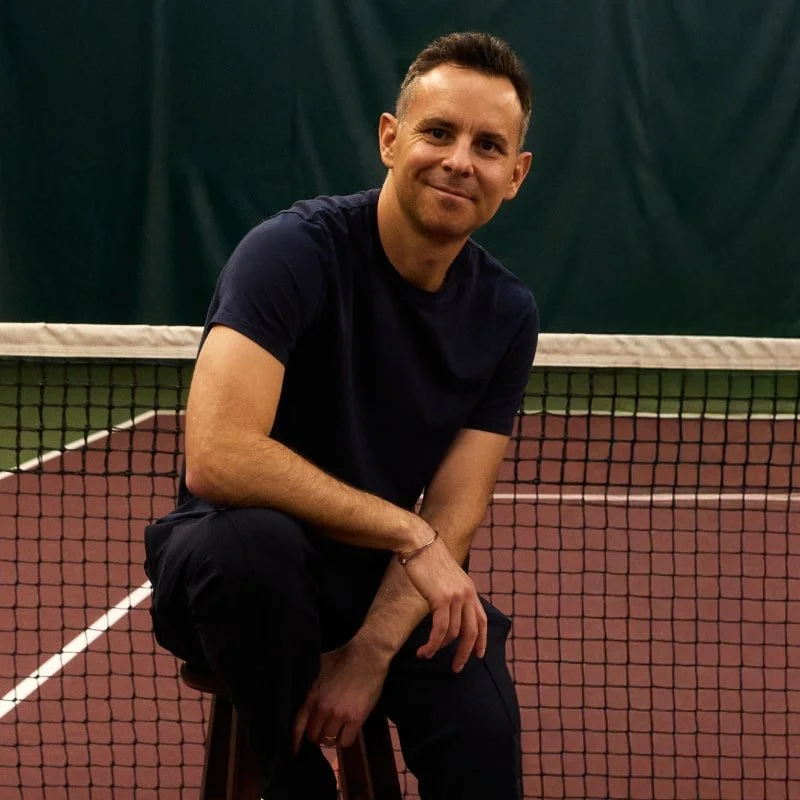 A man with short dark hair, wearing a navy t-shirt and black pants, sitting on a stool next to a tennis net on an indoor tennis court, smiling at the camera.