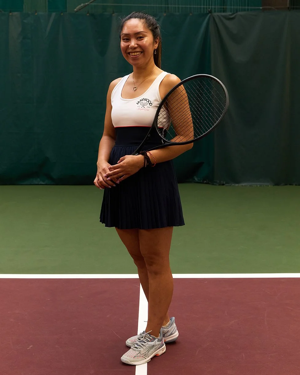 A woman standing on an indoor tennis court holding a tennis racket, smiling and dressed in athletic attire.