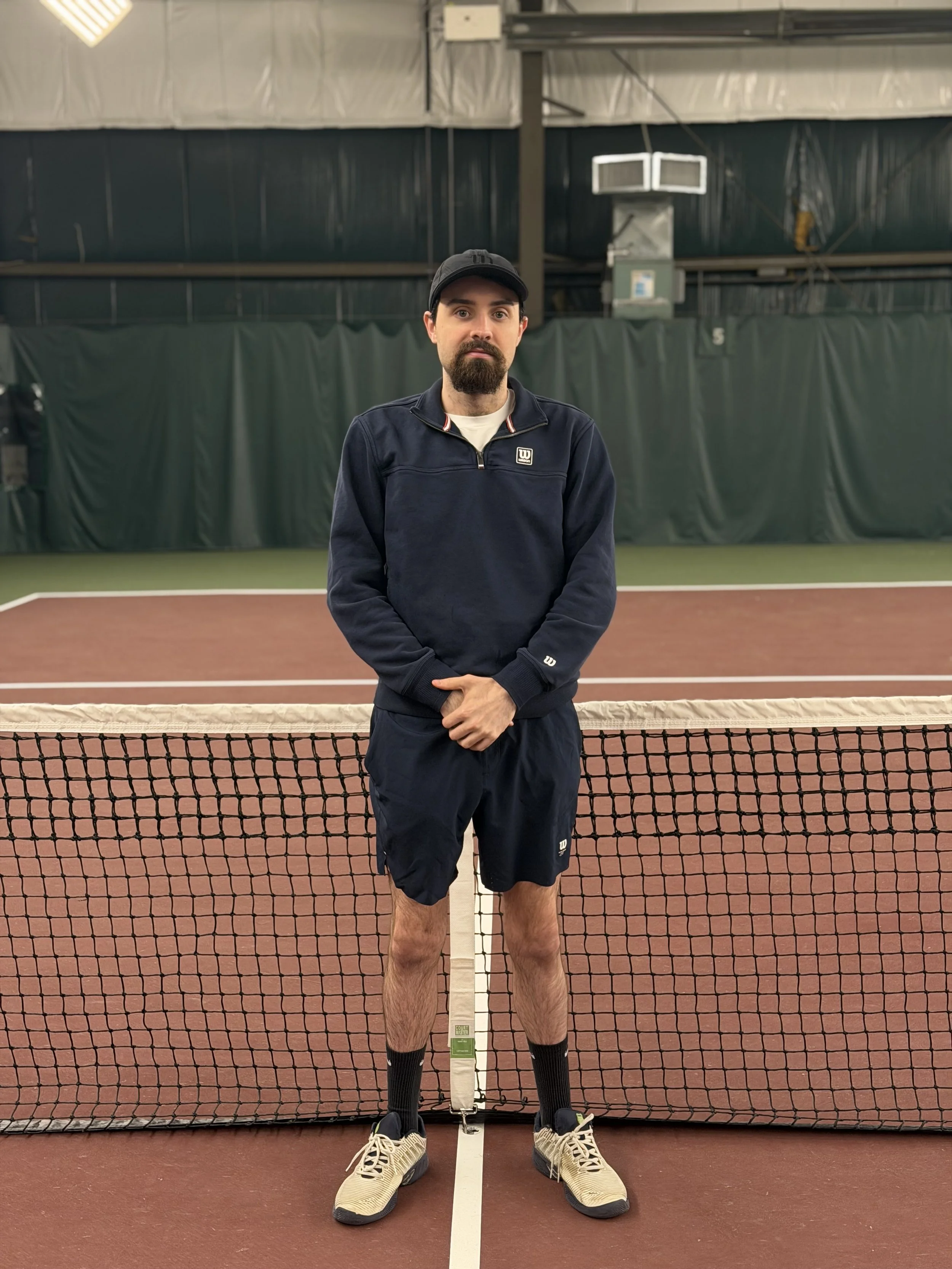 Man standing on an indoor tennis court, positioned behind a tennis net, with a serious expression, wearing a black cap, navy jacket, navy shorts, black socks, and beige sneakers.