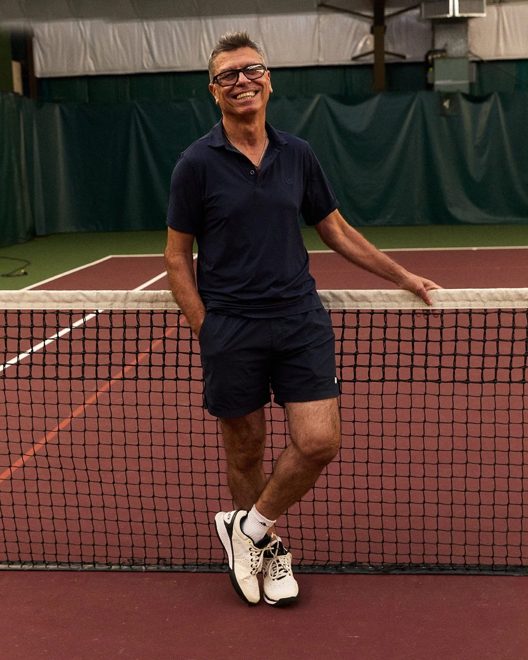 A smiling man wearing glasses, a navy polo shirt, and navy shorts, standing on a tennis court with his arm resting on the net.