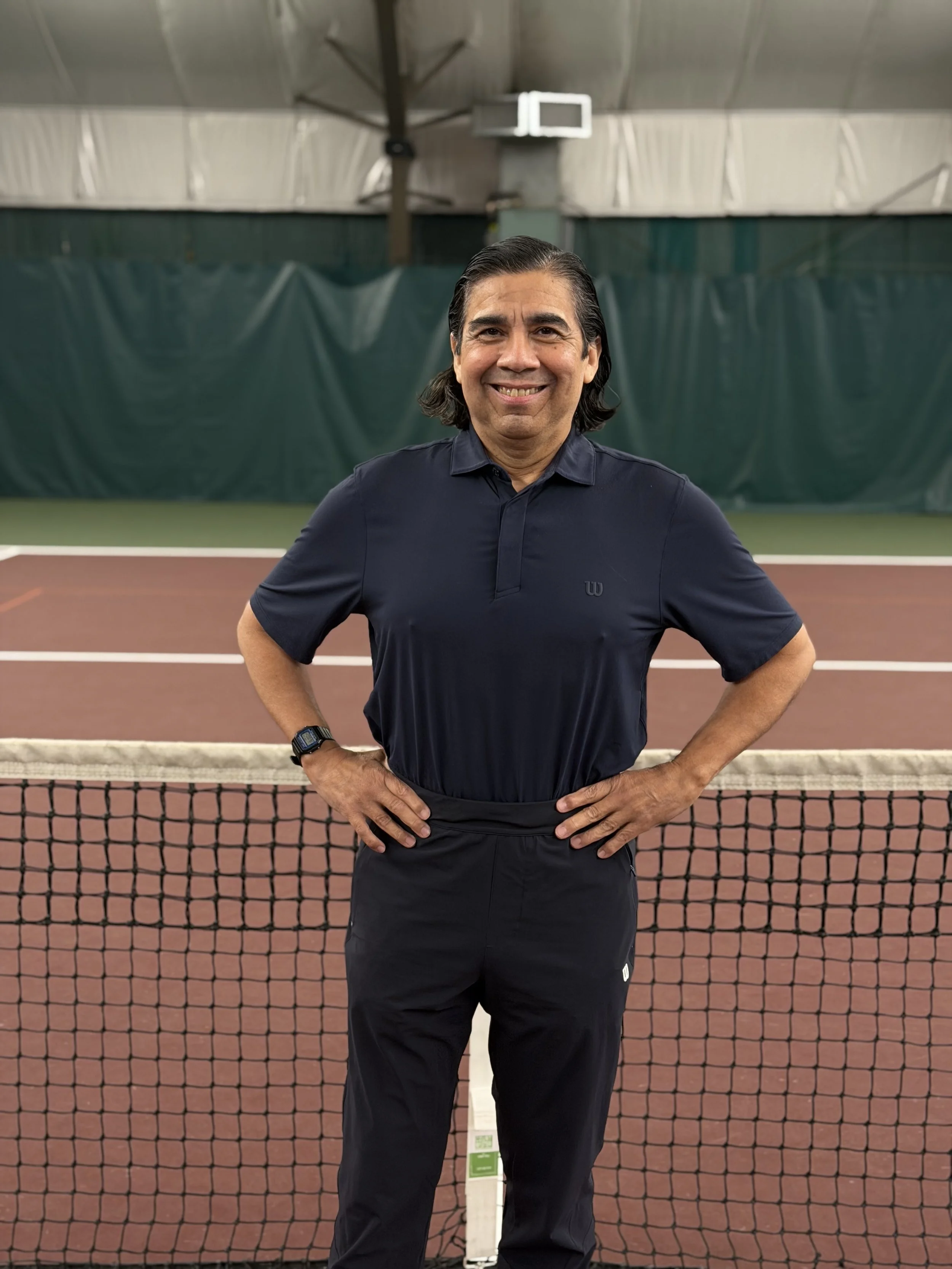 A man standing on an indoor tennis court, smiling, wearing a navy blue polo shirt, black pants, and a watch.