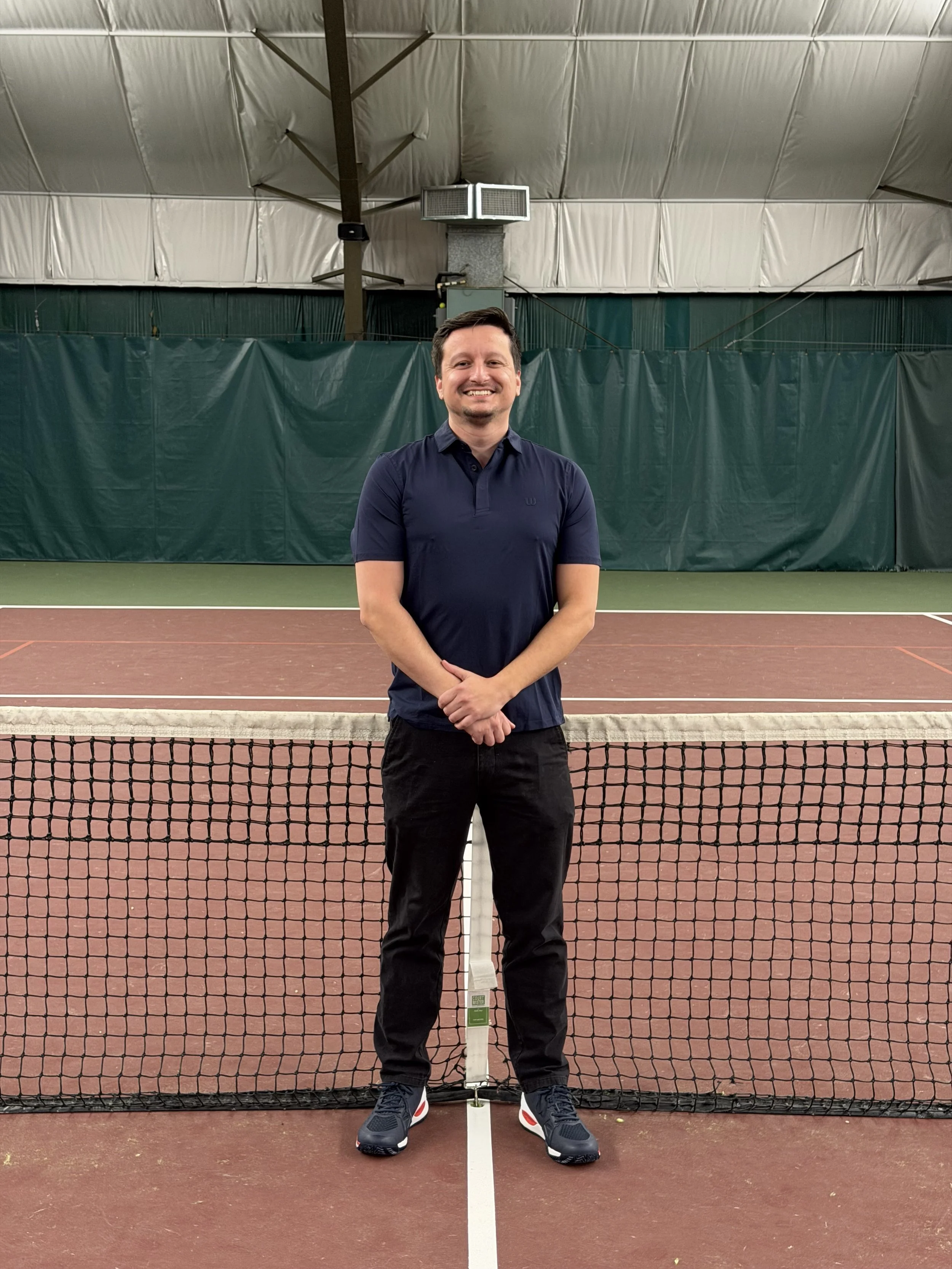 Man standing on an indoor tennis court, smiling, wearing a navy polo shirt, black pants, and sneakers.