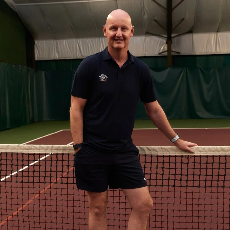 A man standing on an indoor tennis court, smiling, with one hand resting on the net and the other in his pocket, dressed in a dark polo shirt and shorts.