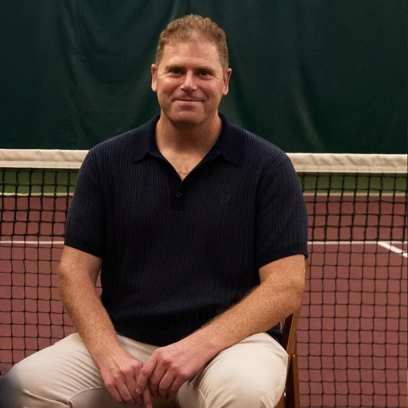 A man sitting on a chair in front of a tennis net inside an indoor tennis court, wearing a dark short-sleeve polo shirt and light-colored pants.