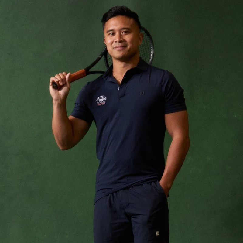 A man in a navy blue polo shirt holding a tennis racket over his shoulder, standing against a green background.