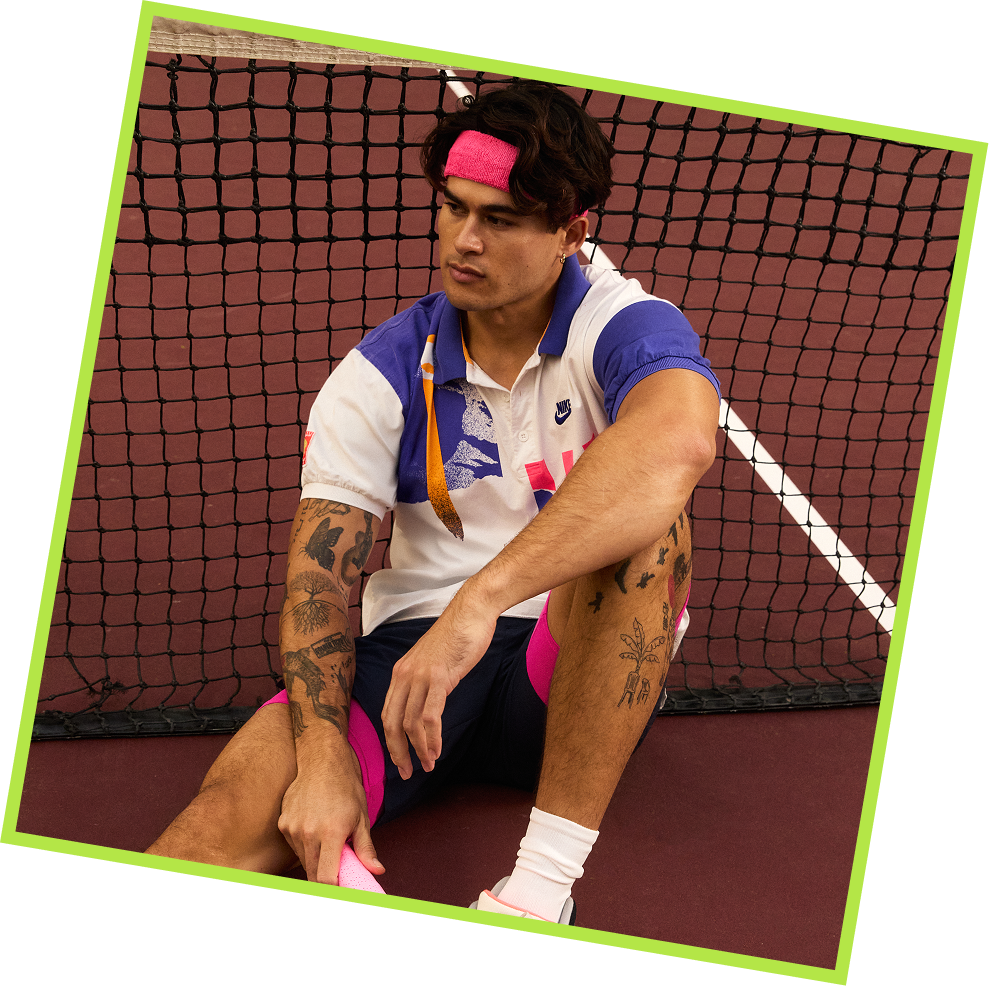 A young man sitting on the tennis court ground in front of a net, wearing a pink headband, a colorful tennis shirt, and shorts, looking pensive.