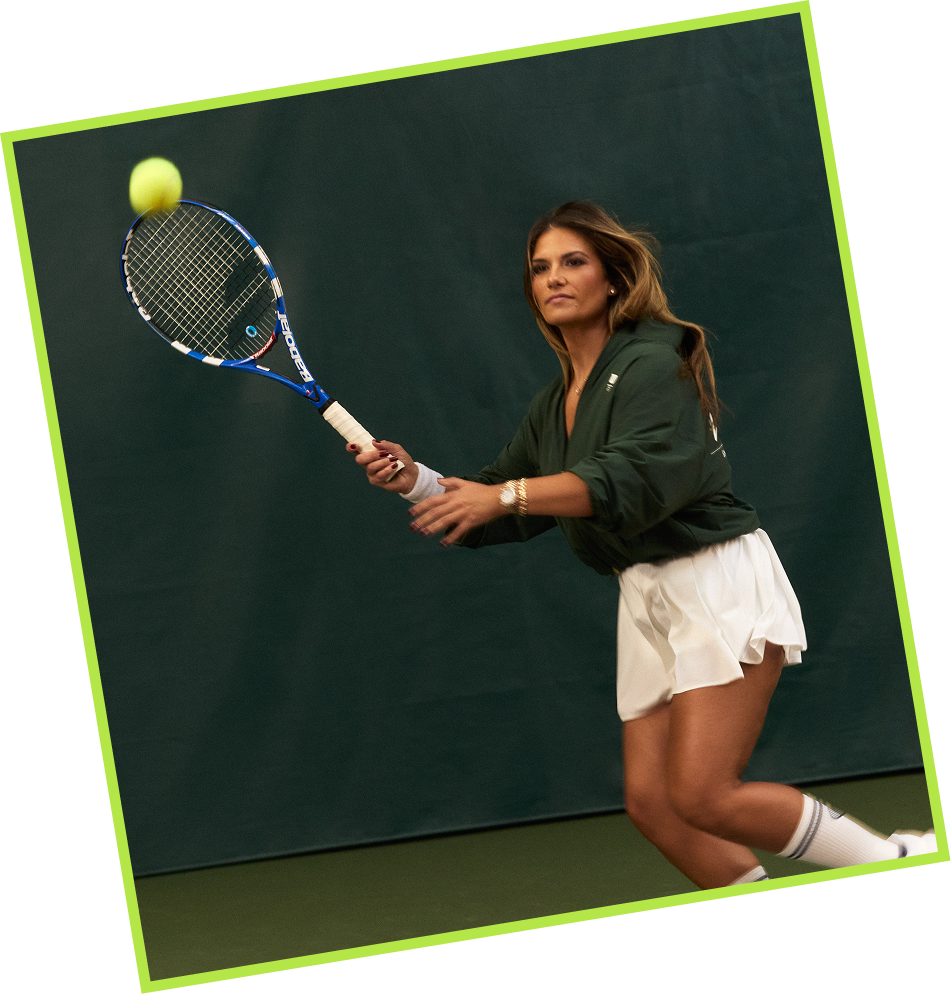 A woman in green sweatshirt and white tennis skirt practicing tennis indoors, hitting a tennis ball with a racket.