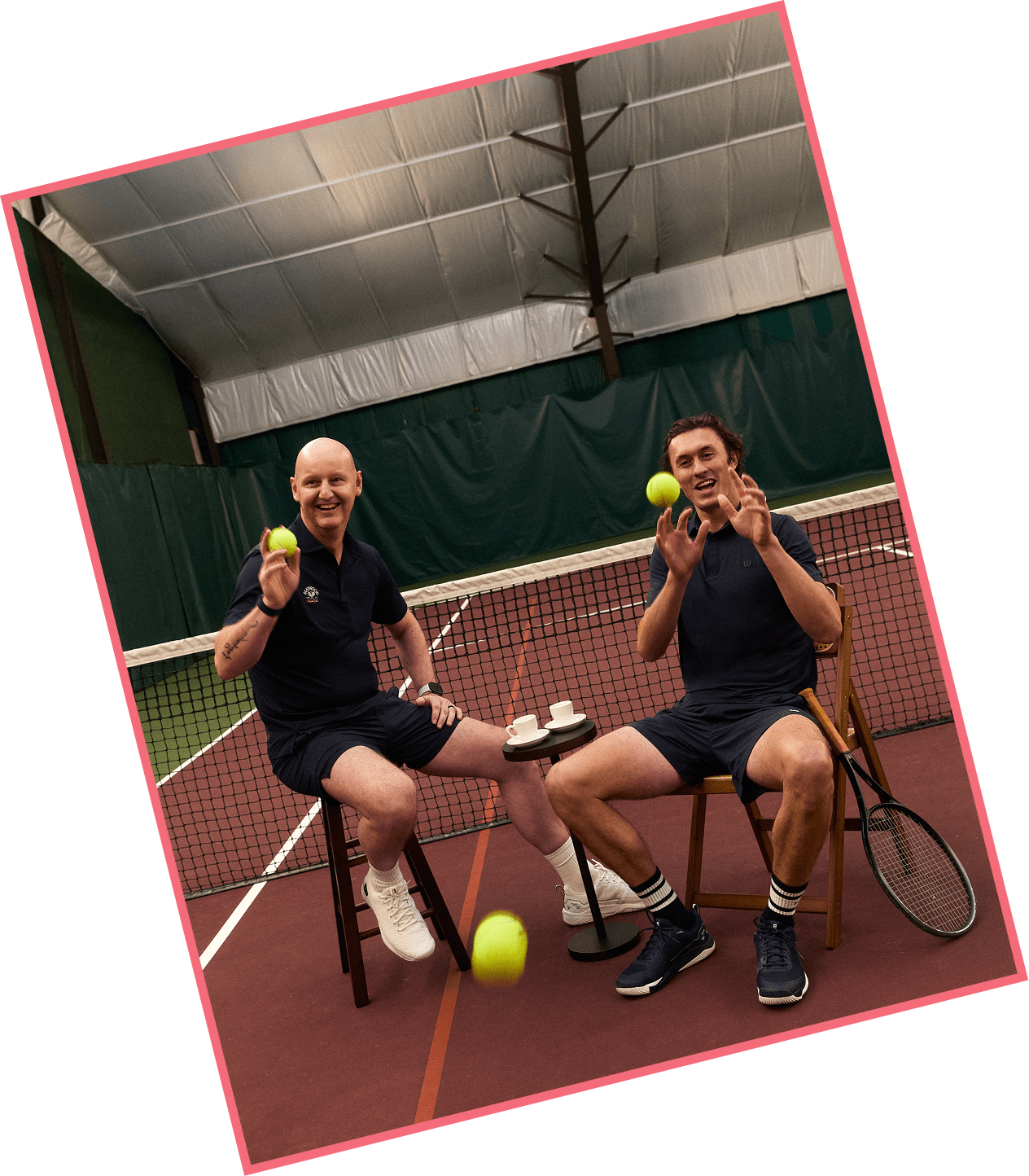 Two men sitting on chairs on a tennis court, holding tennis balls and smiling, with coffee cups and a small table between them.