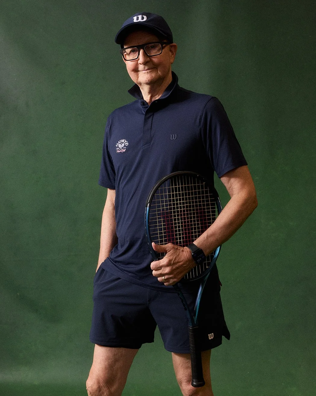An older man dressed in navy blue tennis attire holding a tennis racket, standing against a green background.