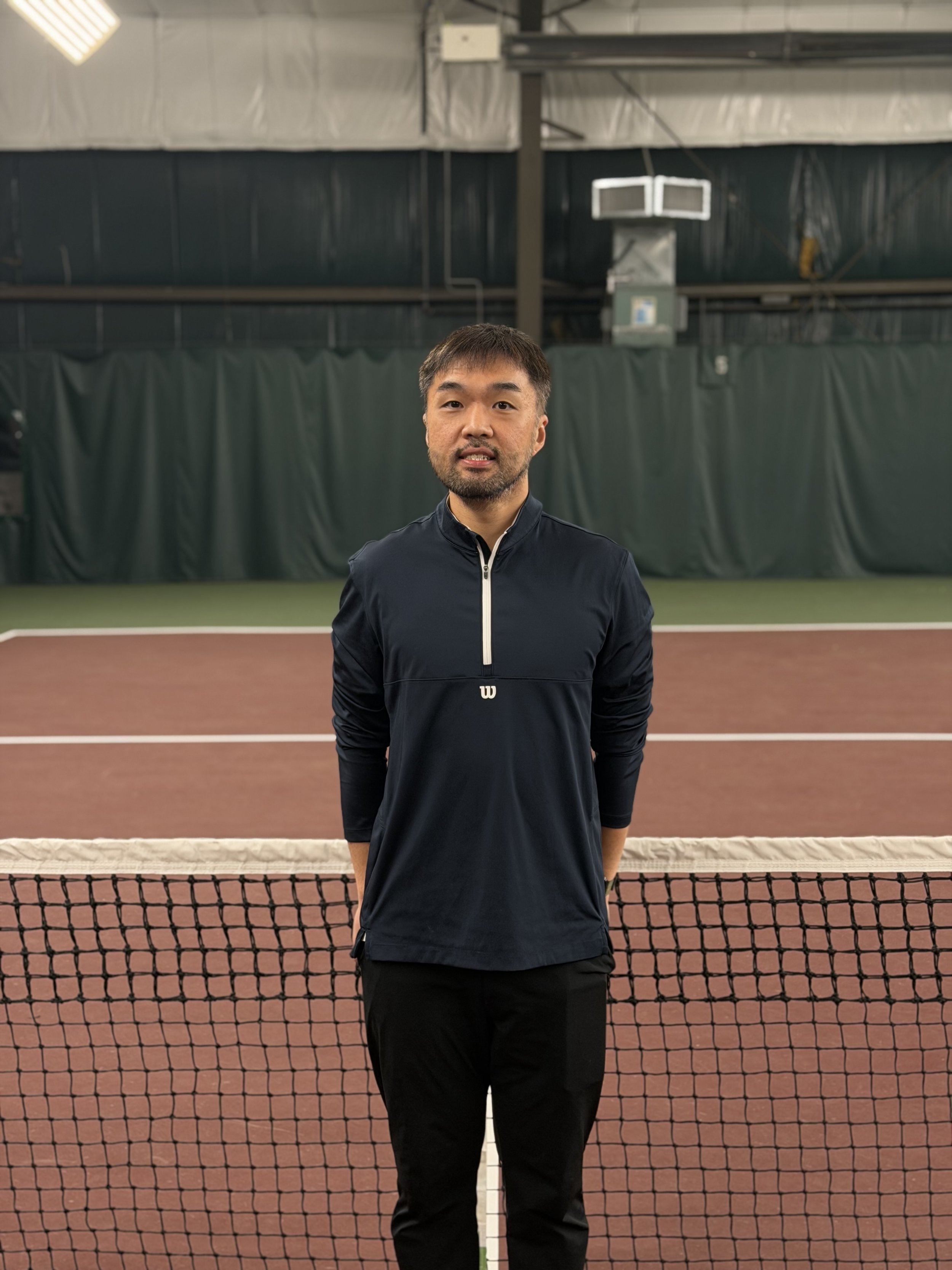A man standing on an indoor tennis court behind the net, wearing a navy blue sports jacket and black pants.