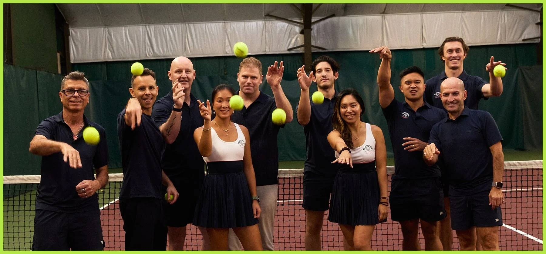 Group of people on an indoor tennis court playing with tennis balls, some midair, smiling and posing for the camera.