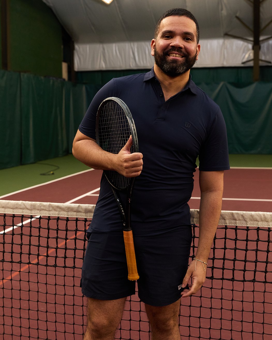 A man with dark hair and a beard smiling on an indoor tennis court, holding a tennis racket, wearing a dark blue polo shirt and shorts.