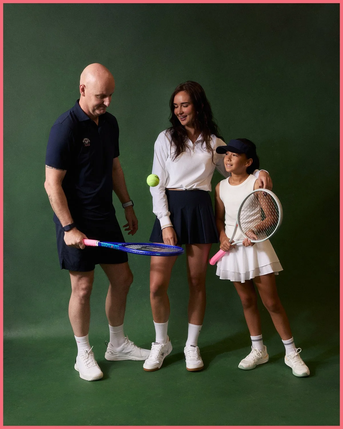 A man, woman, and young girl standing together against a green background, holding tennis rackets and a tennis ball, dressed in tennis attire.