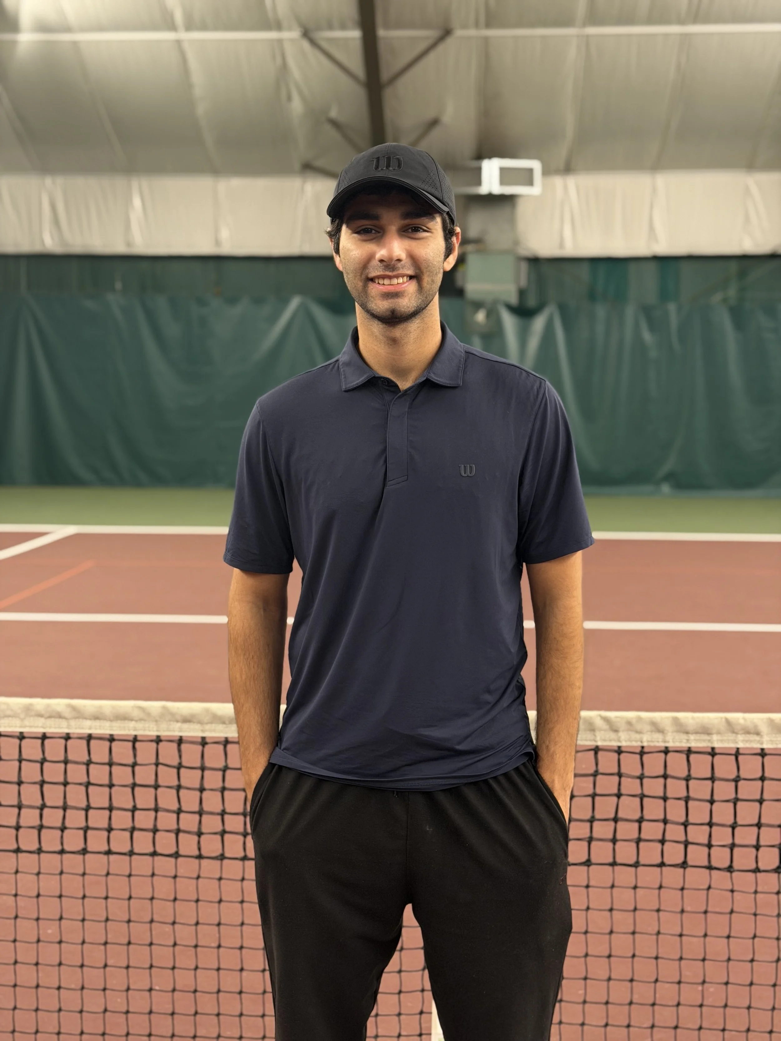 A young man standing on an indoor tennis court, wearing a dark polo shirt, black shorts, and a black cap, with a tennis net in front of him.