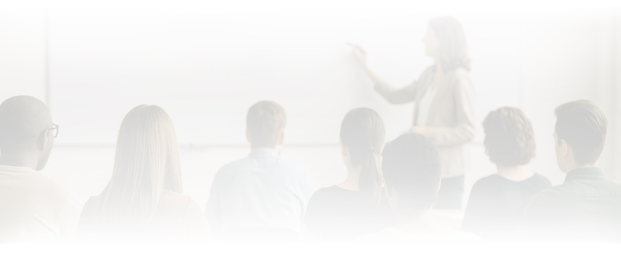 A woman is standing in front of a group of people in a classroom, giving a presentation or lecture. The audience members are seated and facing her.
