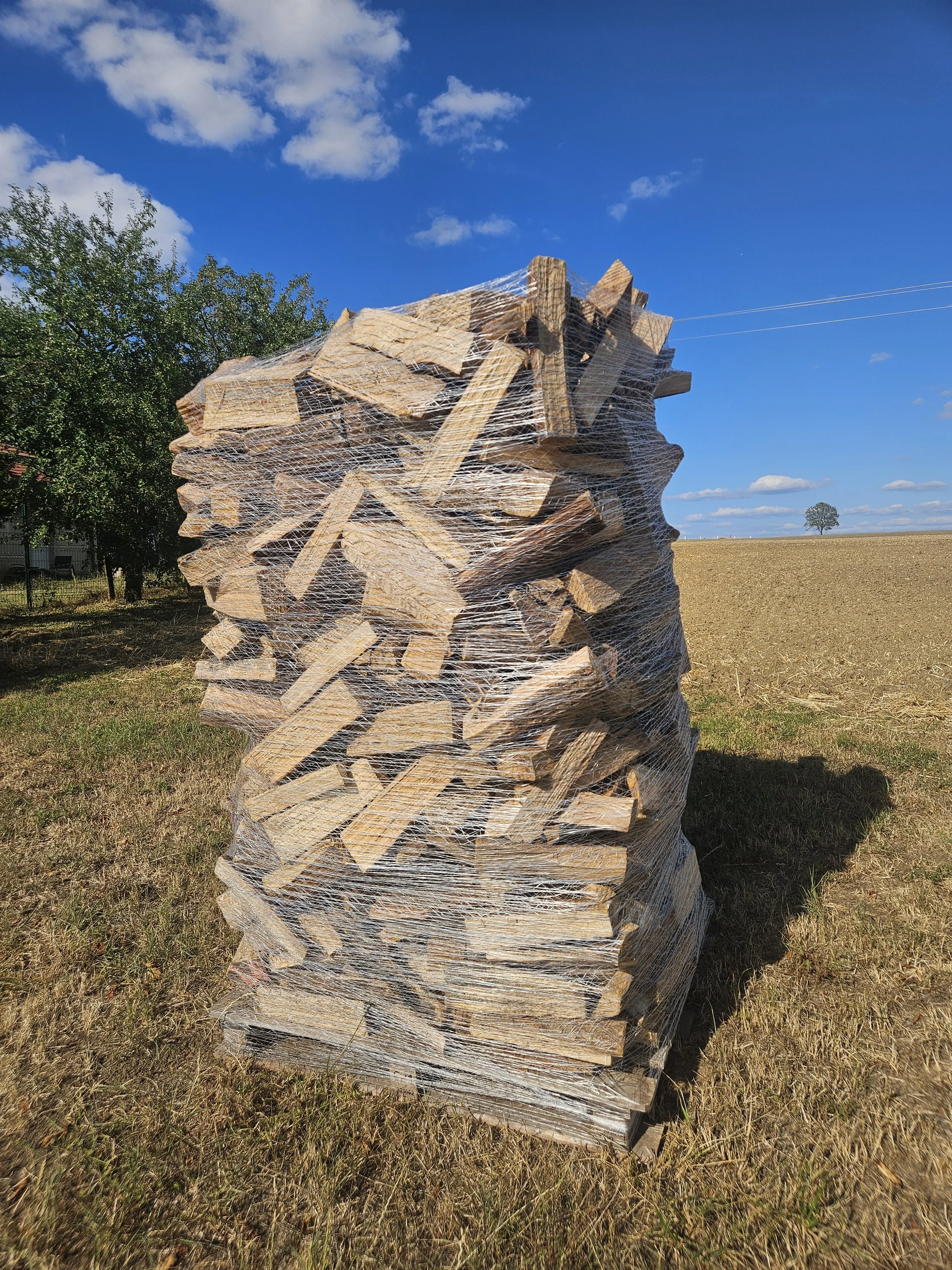 Tas de bois empilés et emballés avec du filet en plastique dans un champ rural sous un ciel bleu avec quelques nuages.