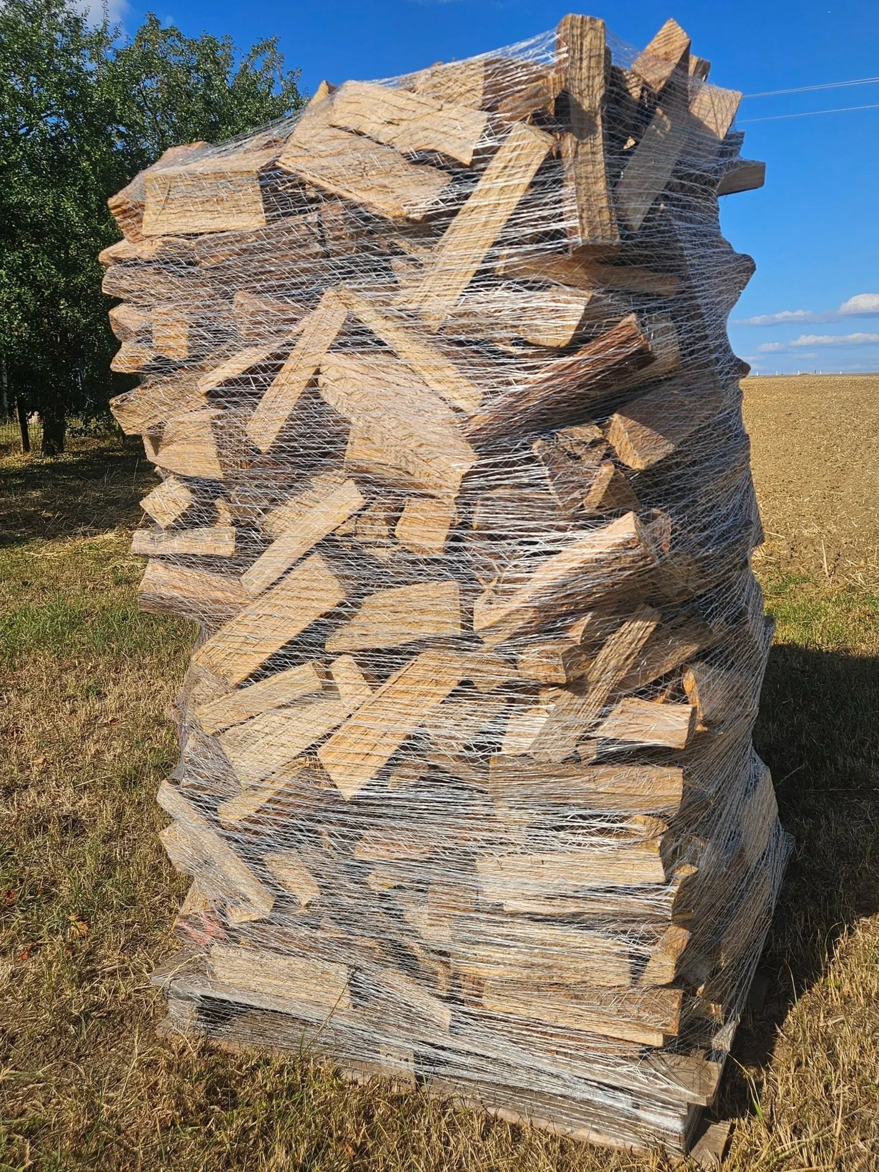 pile de bois coupé enveloppée dans du film plastique, située dans un champ en plein air, avec un ciel bleu clair en arrière-plan.