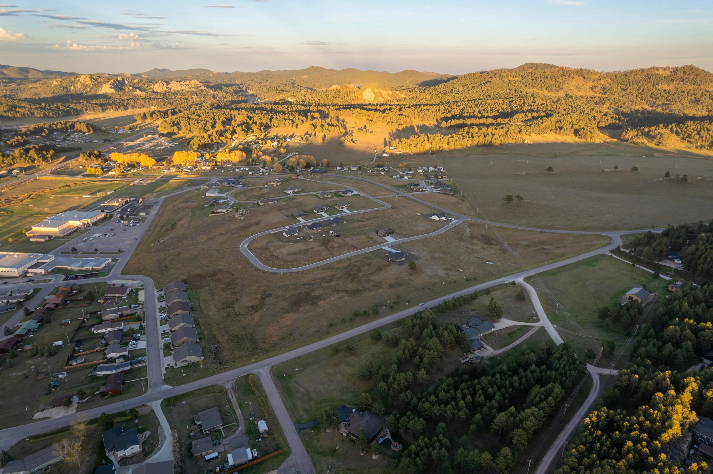 Aerial view of a semi-rural area with houses, roads, open fields, and a wooded area with mountains in the background at sunset.