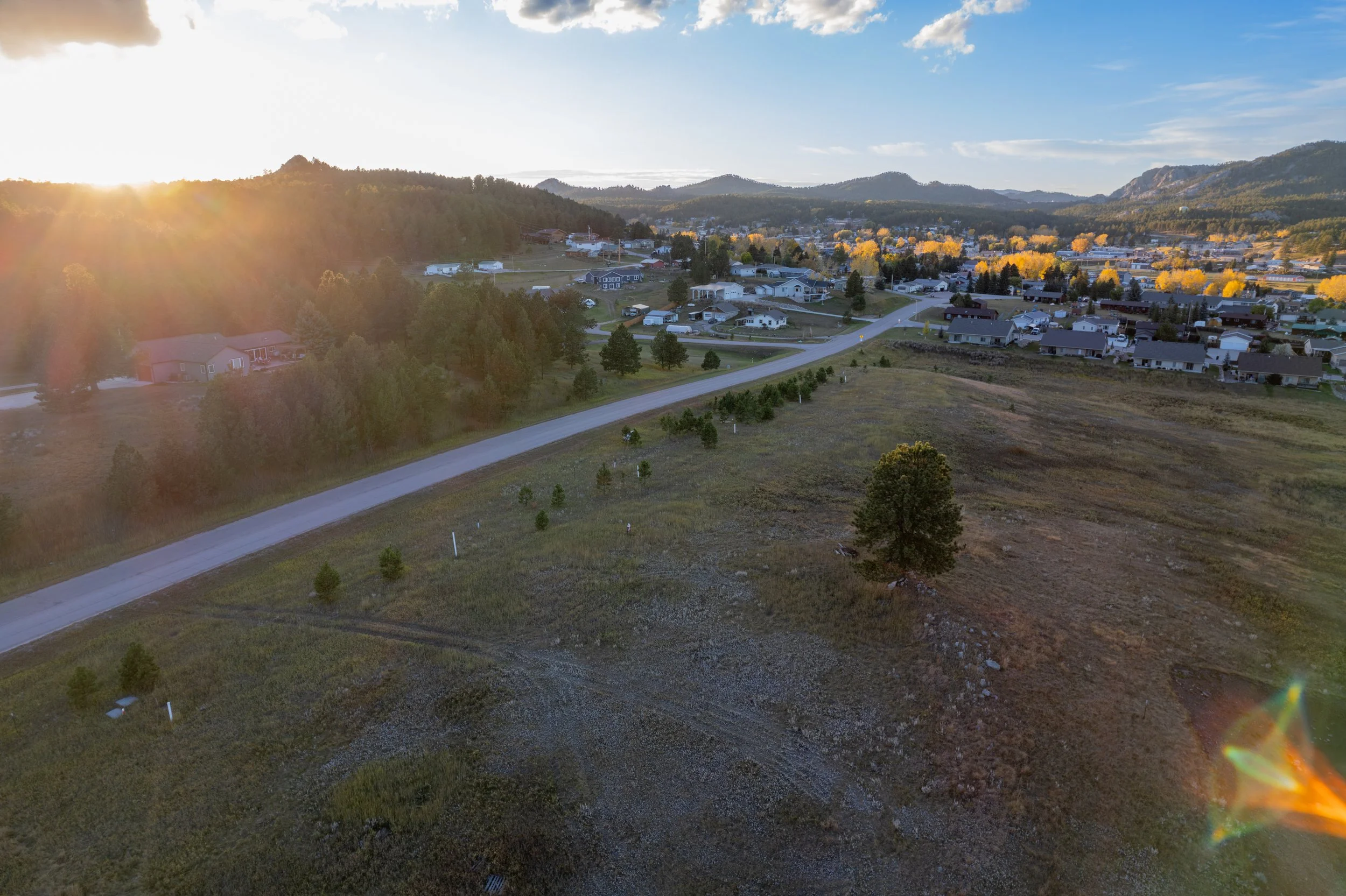 Aerial view of a suburban neighborhood at sunset with a dirt road, houses, trees, and distant mountains.