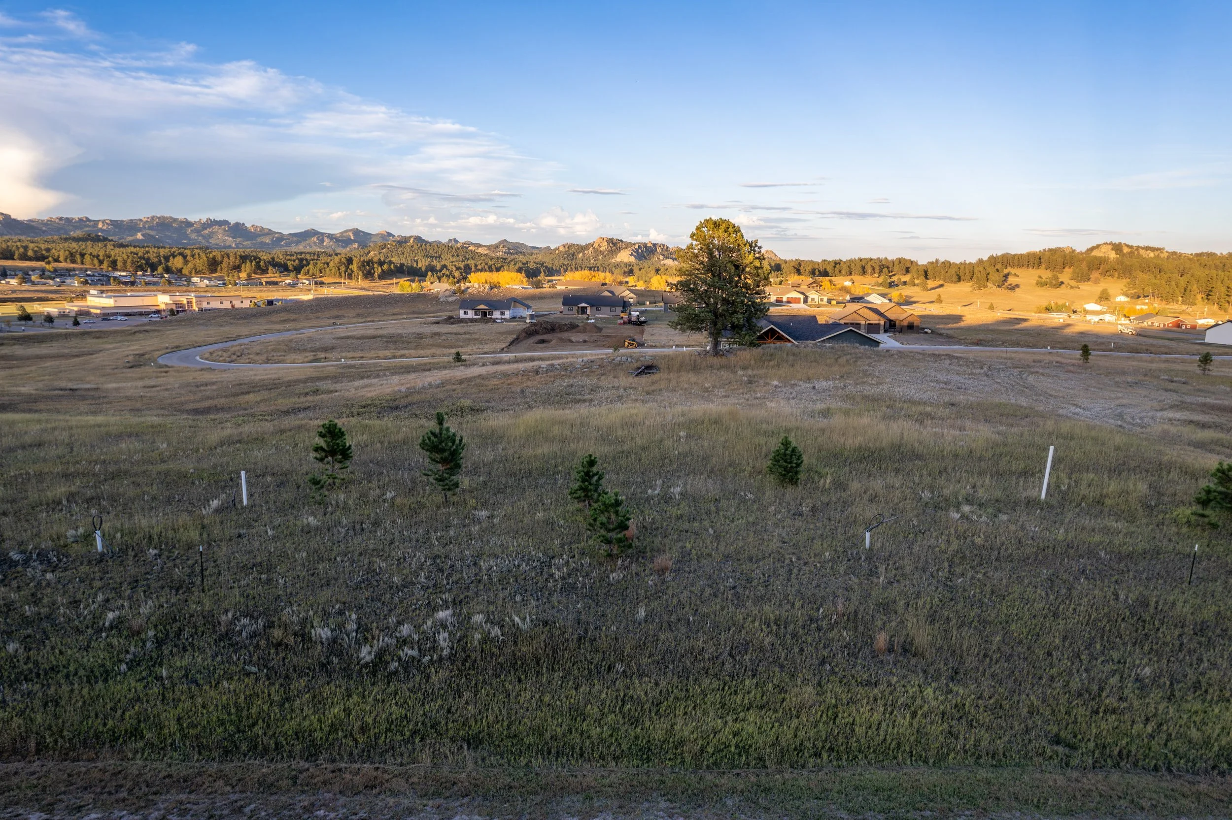 Scenic view of a rolling countryside with small trees, a winding road, scattered houses, and mountains in the background under a partly cloudy sky during sunset.