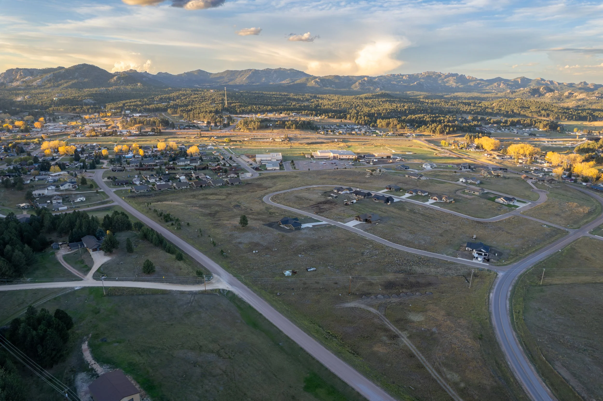 Aerial view of a suburban neighborhood with houses and roads, surrounded by open fields and distant mountains under a partly cloudy sky.