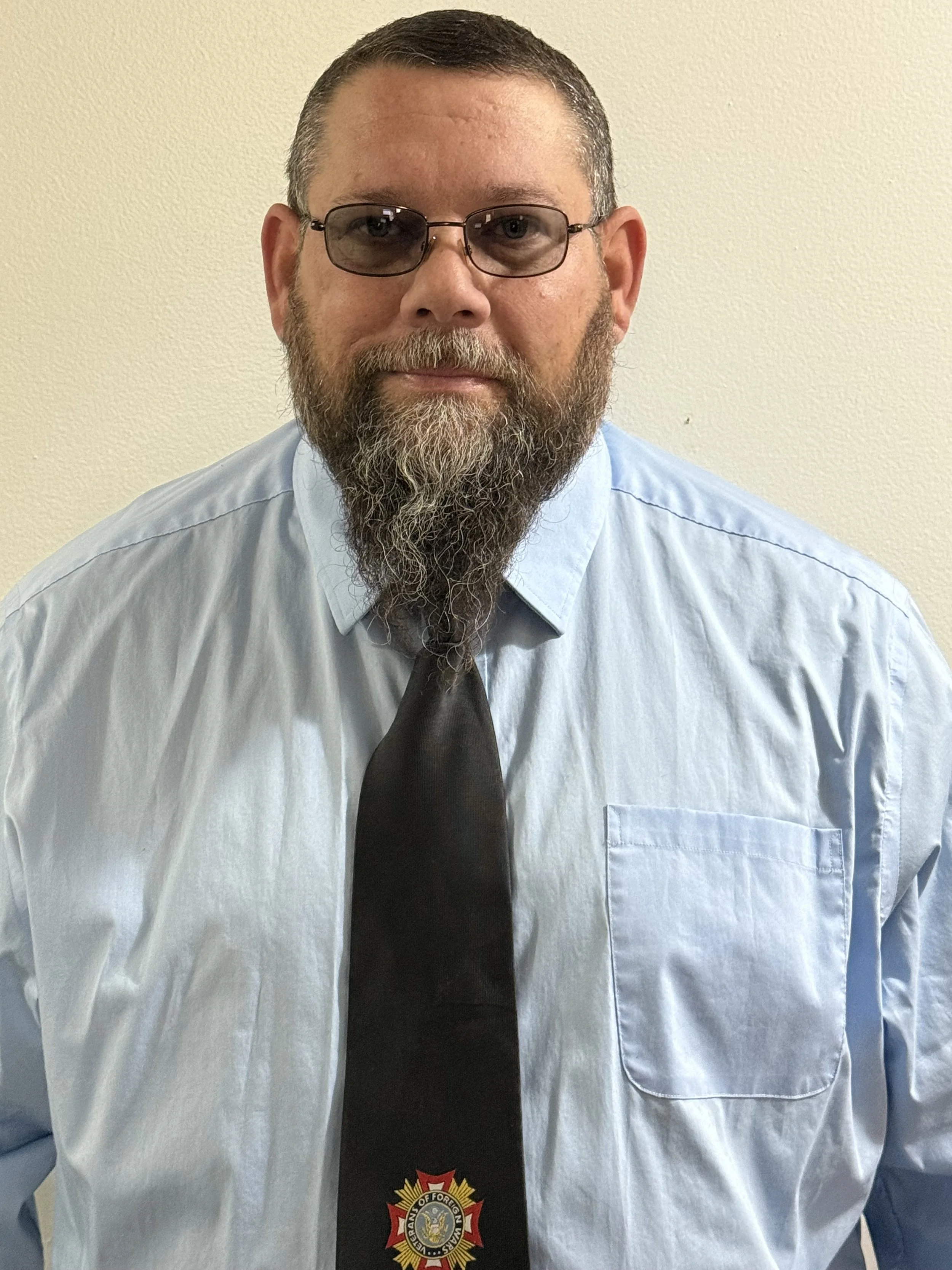 A man with glasses, a beard, and a mustache, wearing a light blue dress shirt and a black tie with a fire department emblem, standing against a plain wall.