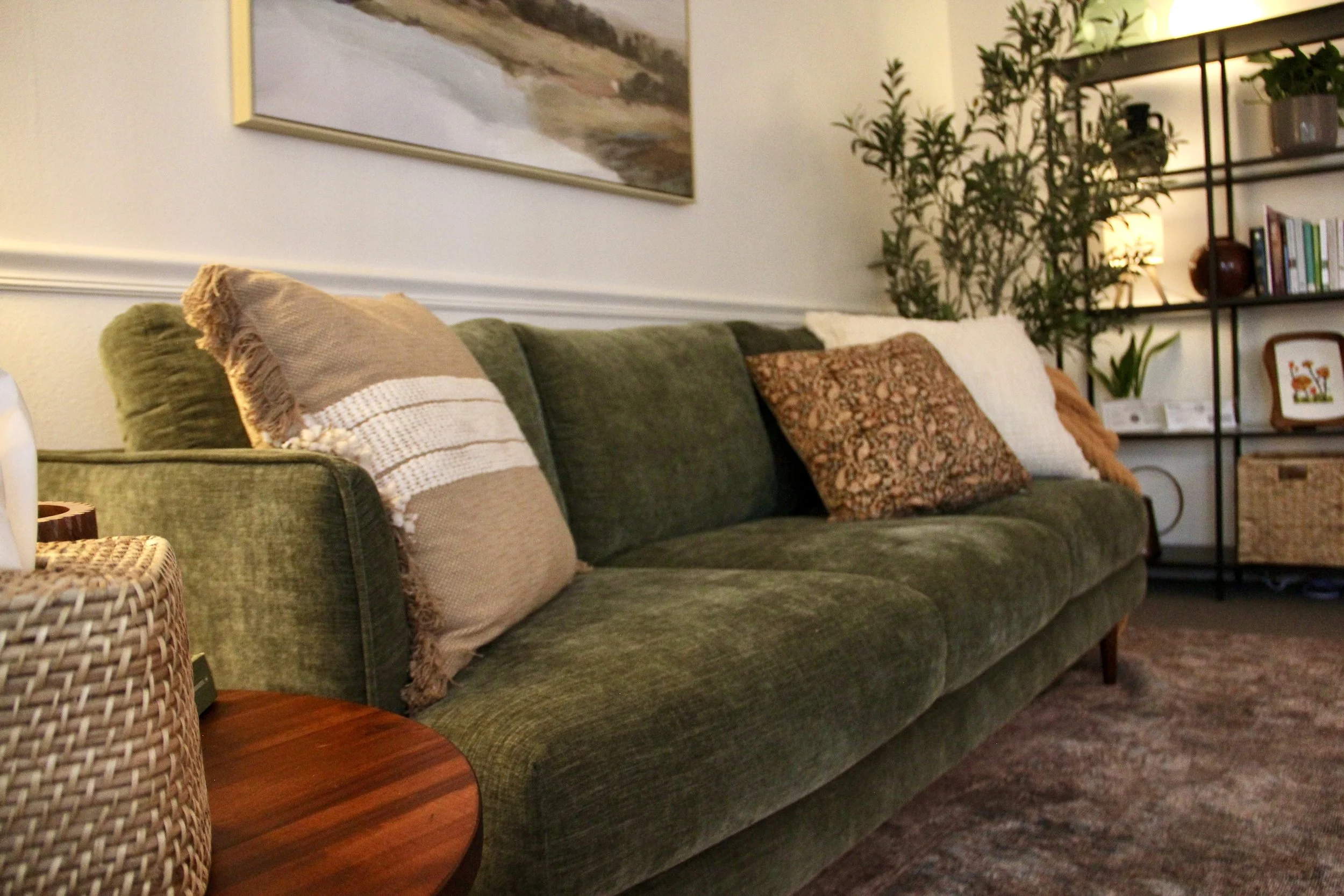 A green velvet sofa with four decorative pillows of different colors and patterns, positioned in a cozy living room with a framed landscape painting, a bookshelf with plants and books, a woven basket, and a brown wooden coffee table.