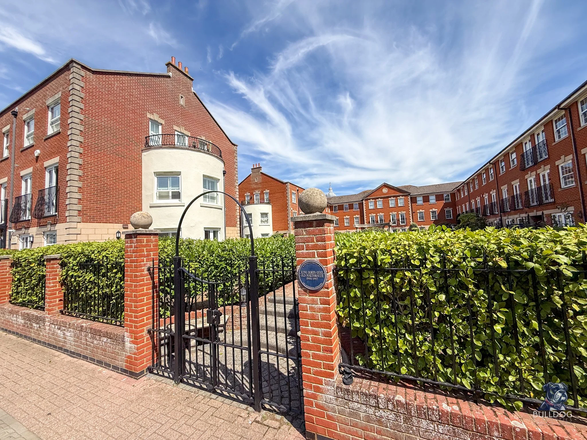 Brick buildings with white accents, balconies, and green shrubbery enclosed by a black iron gate and brick pillars, under a partly cloudy sky.