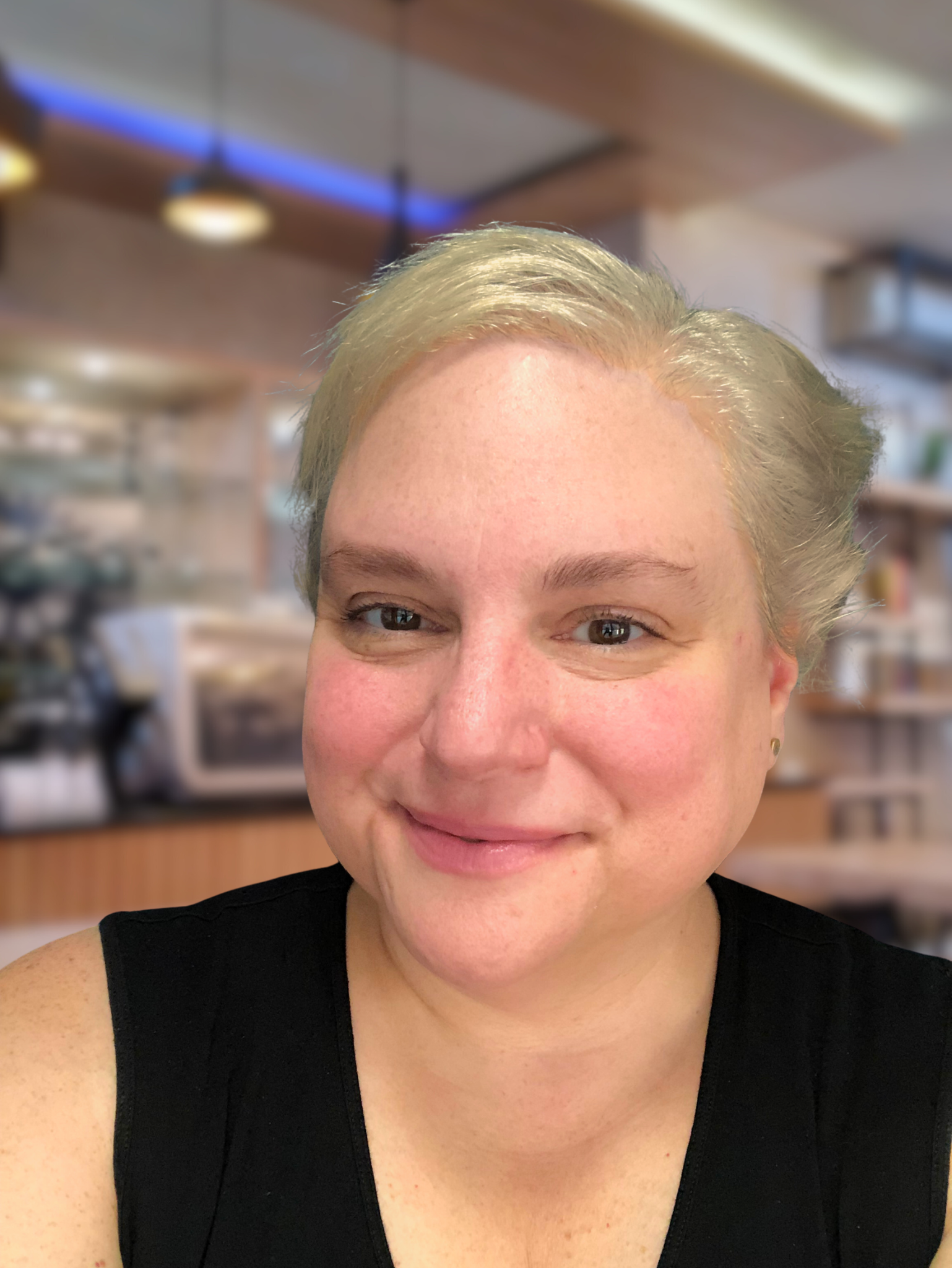 A woman with short blonde hair smiling in a cafe with warm lighting and wooden decor.