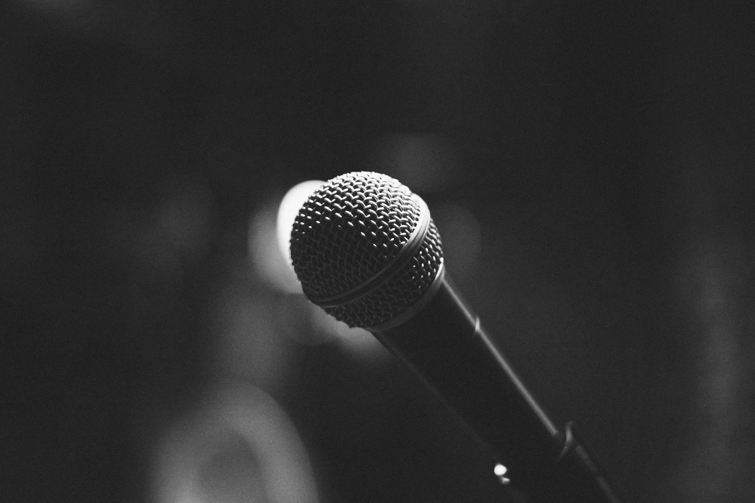A close-up black and white photograph of a microphone on a stand against a dark background.