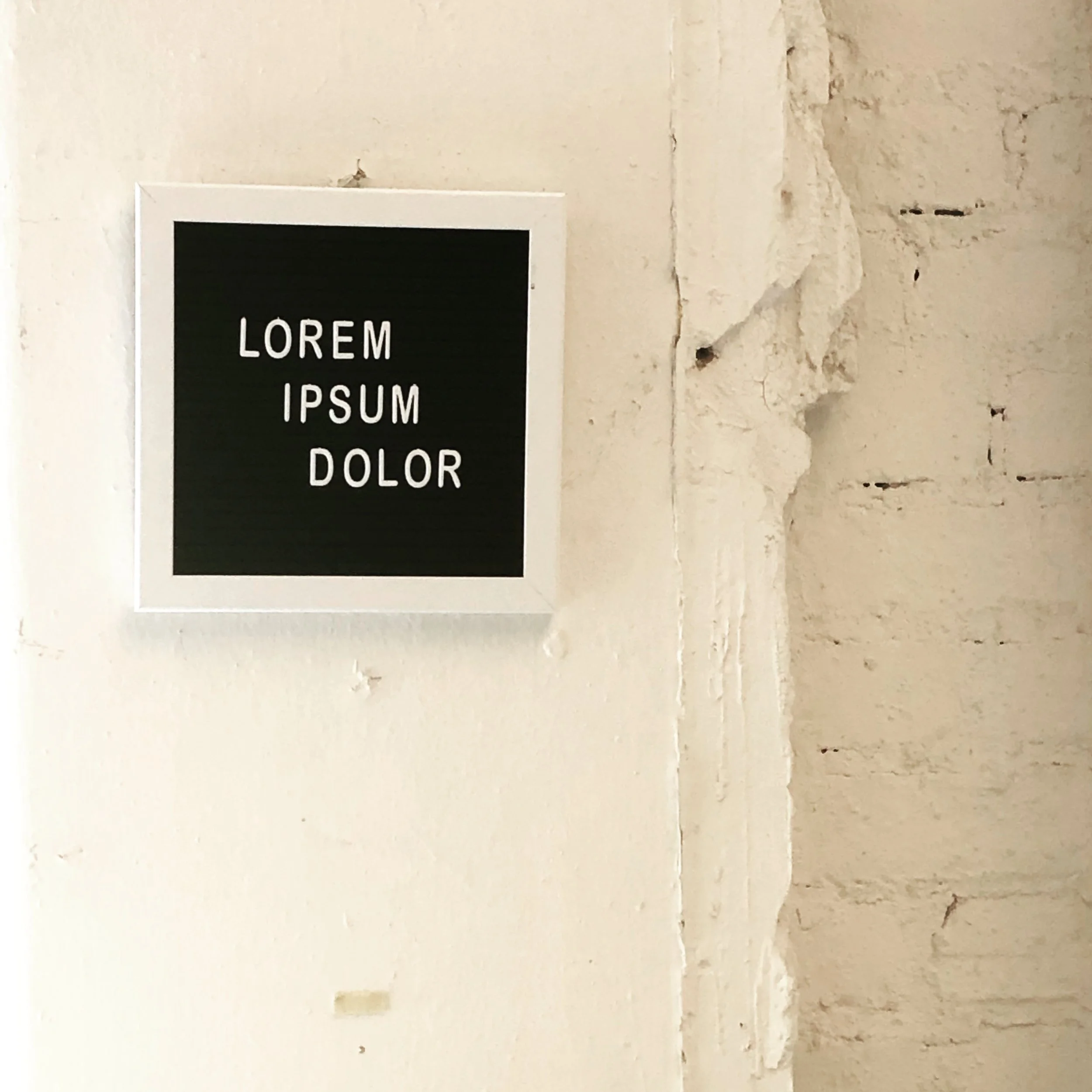 A wall with a small white-bordered black square sign displaying "LOREM IPSUM DOLOR" in white text, adjacent to a rough, partially damaged white brick wall.
