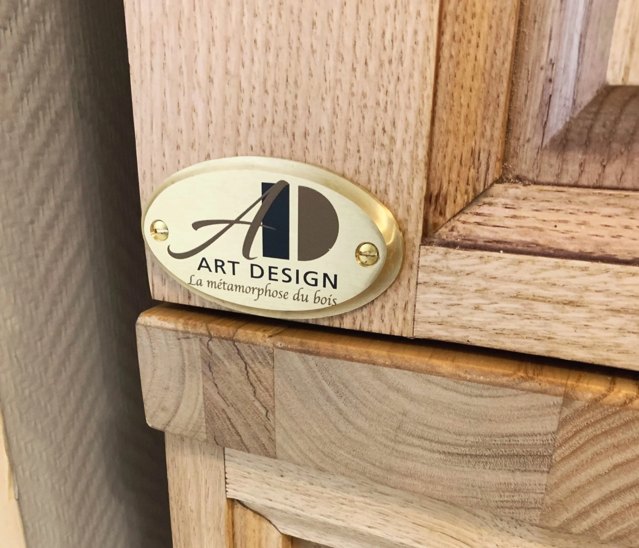 Plaque de métal oval avec inscription, fixée sur un meuble en bois. L'inscription indique 'Art Design' et 'La métamorphose du bois'.