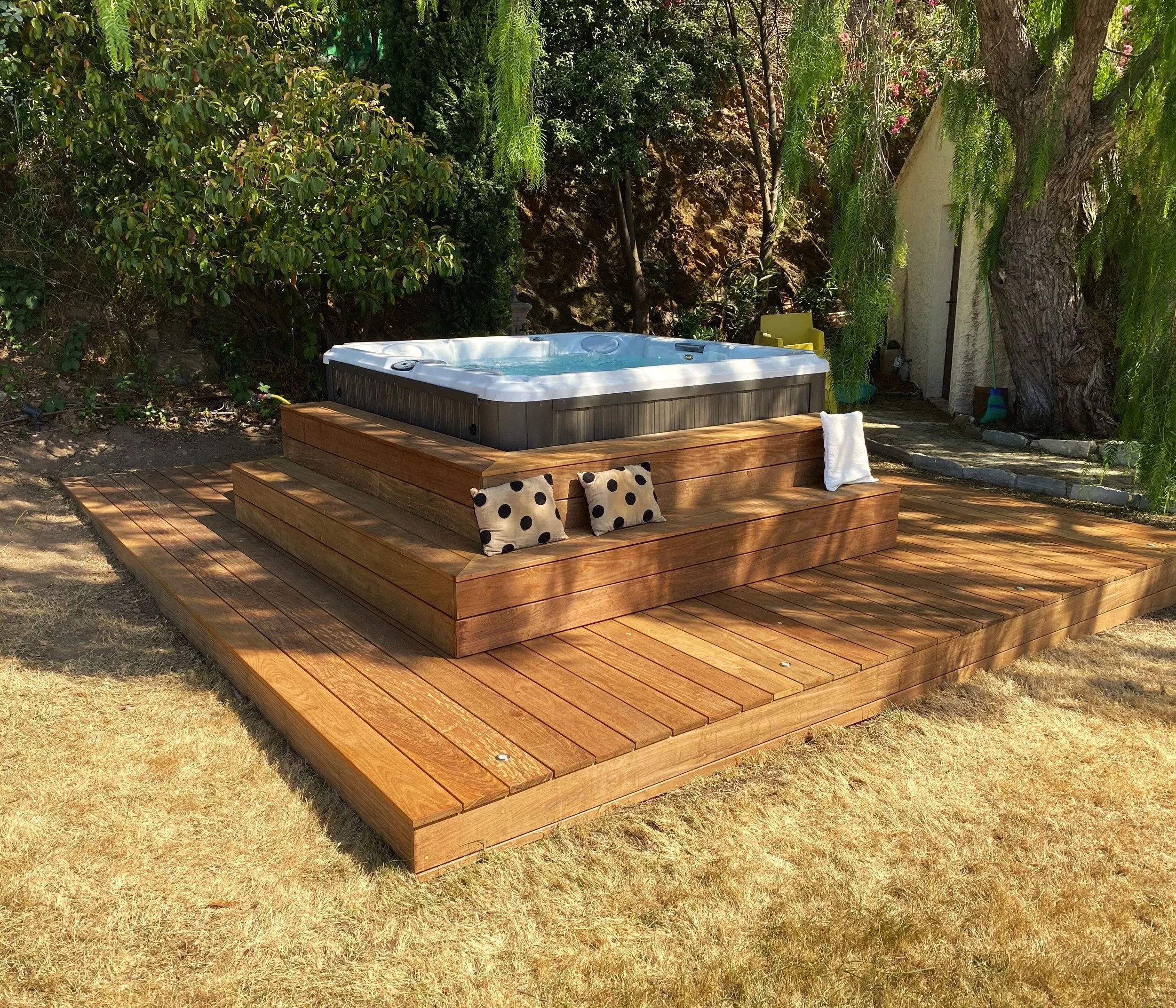 Jacuzzi en bois dans un jardin avec coussins noirs à pois blancs et serviette blanche, entouré d'arbres et de végétation.