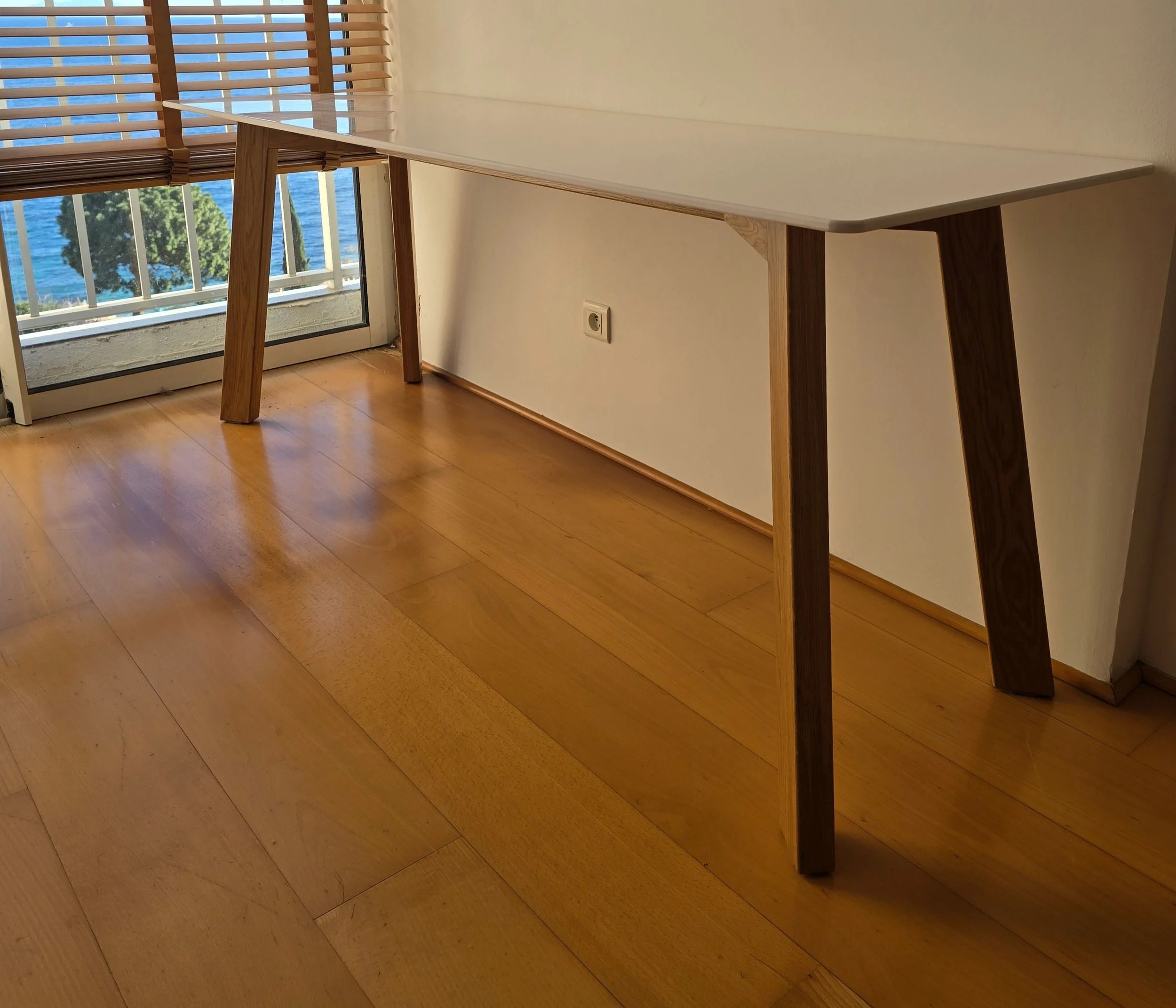 Bureau en bois clair près d'une fenêtre avec volets en bois, vue sur la mer et des arbres à l'extérieur.