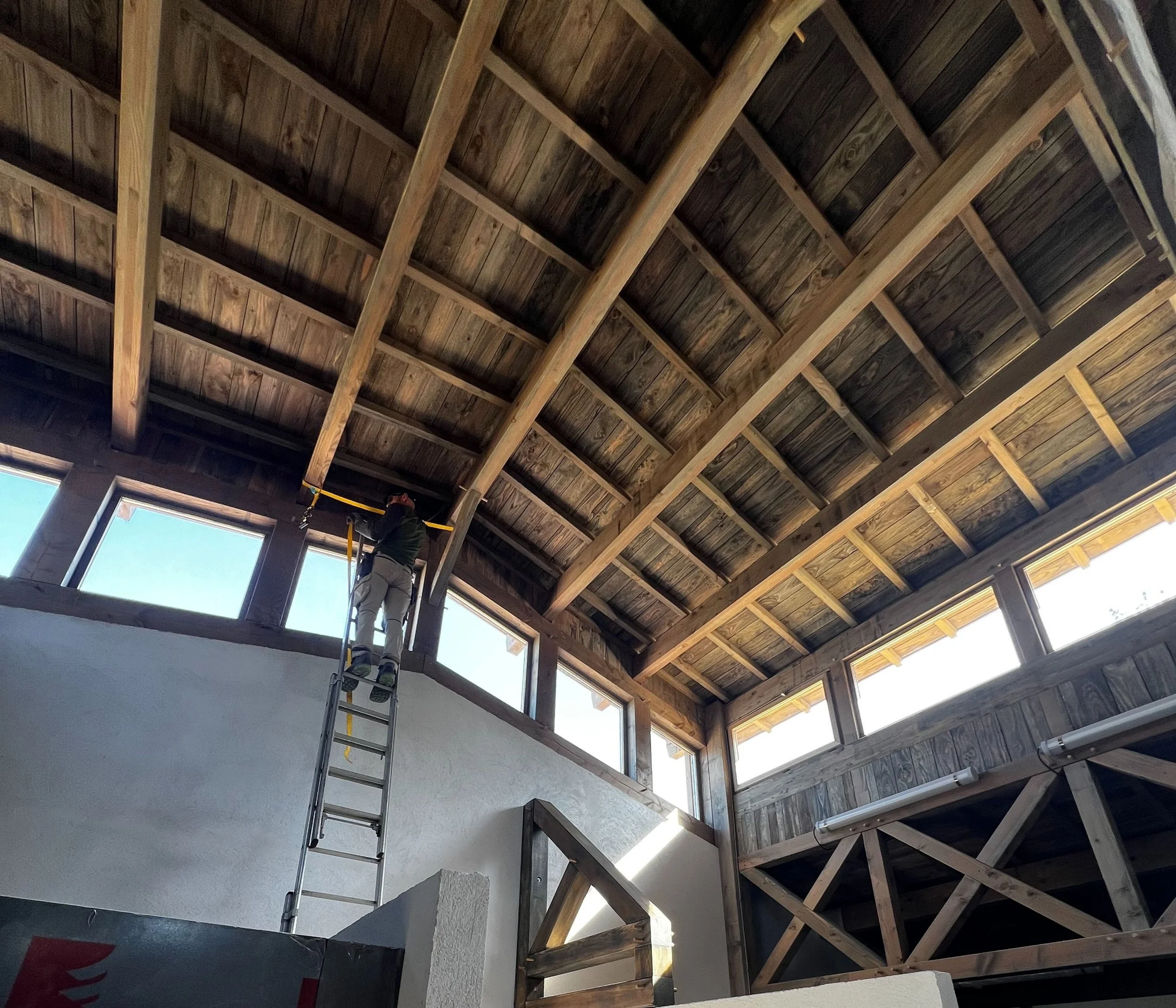 Un homme debout sur une échelle en train d'installer ou de réparer des fenêtres dans une pièce à plafond en bois avec beaucoup de lumière naturelle.