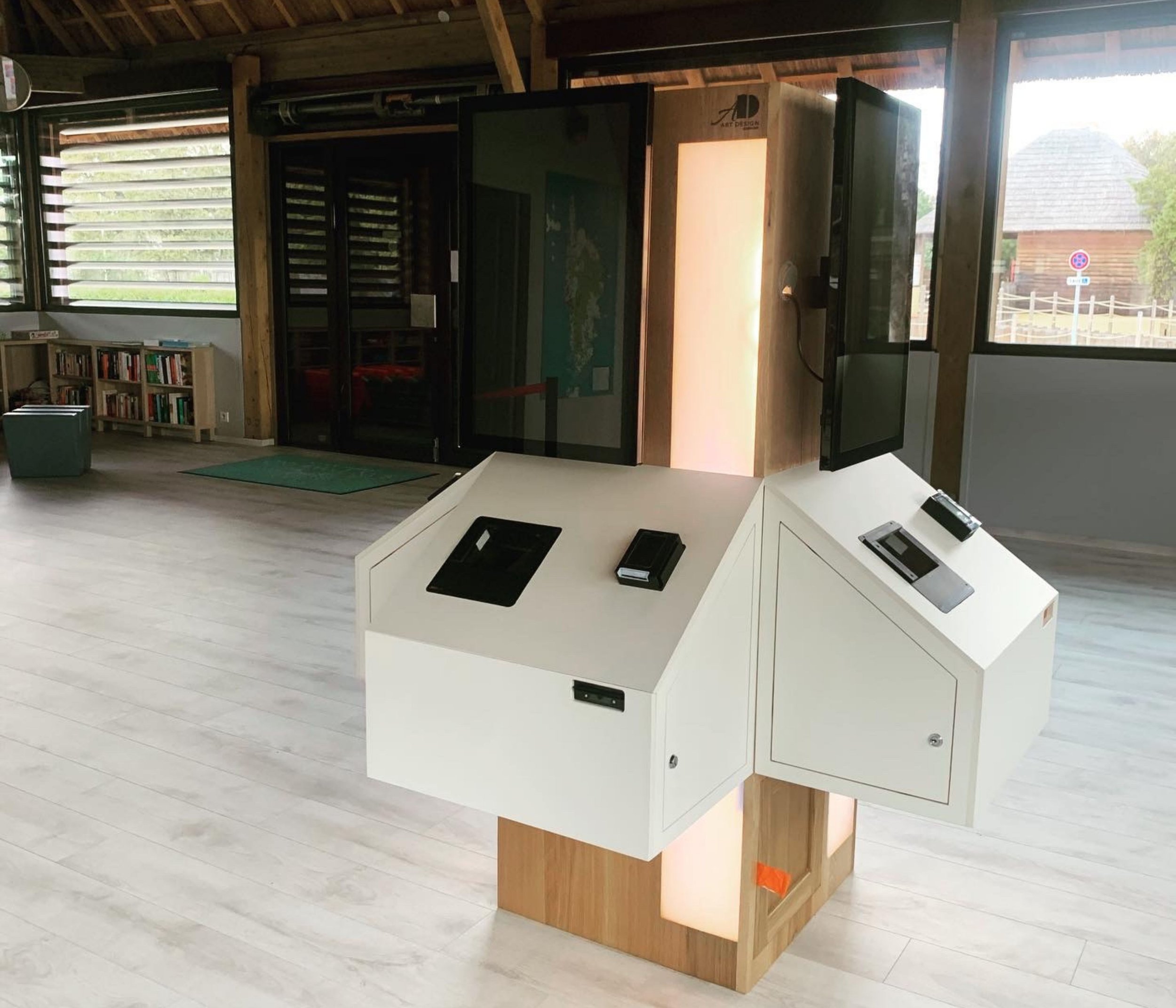 Une installation numérique interactive dans un espace intérieur moderne, avec un dispositif blanc et plusieurs écrans, devant des fenêtres laissant entrer beaucoup de lumière naturelle.