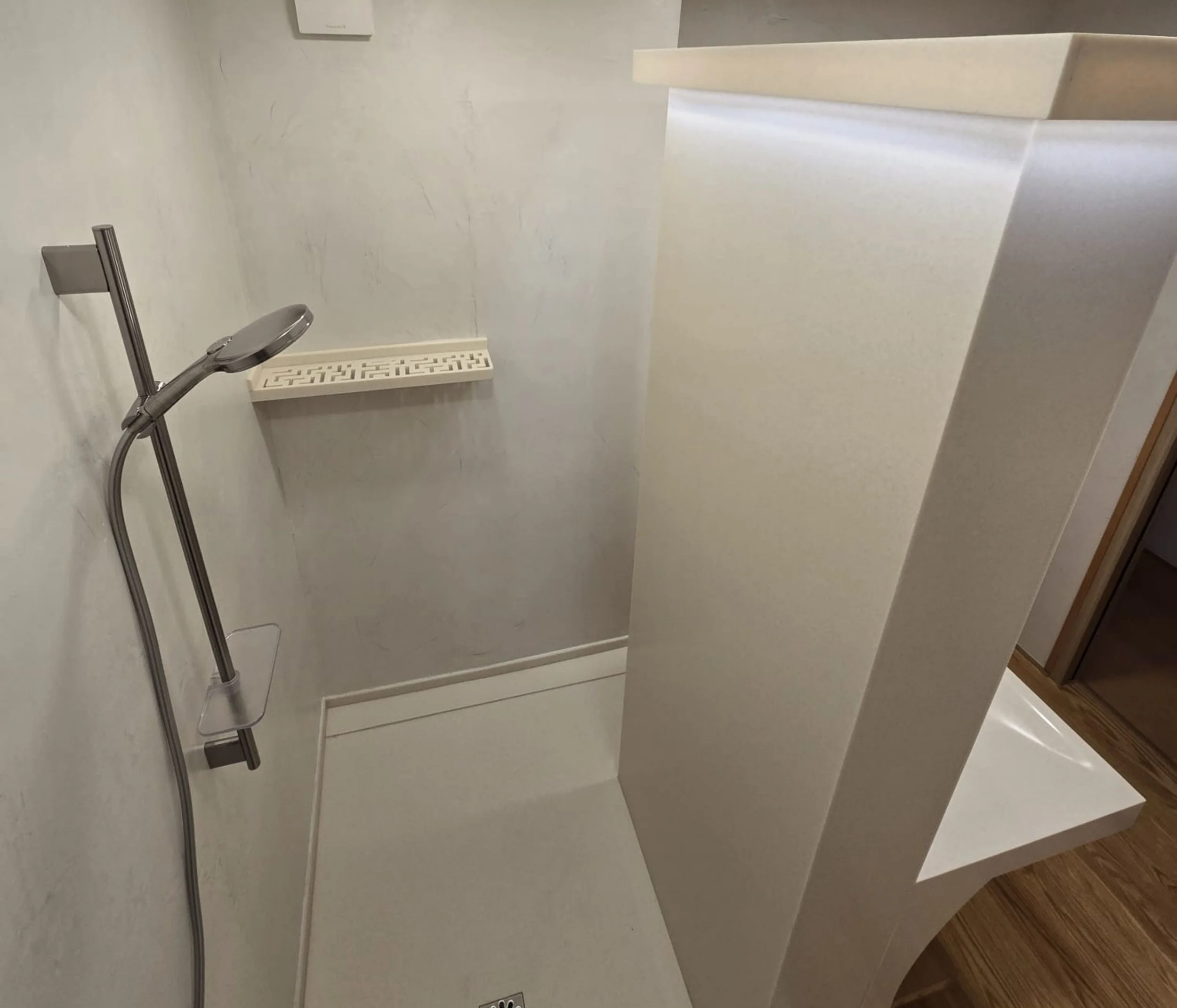 Une douche moderne compacte avec une douchette à main, un petit banc blanc intégré, et un drain au sol dans une salle de bain aux murs clairs et à la décoration minimaliste.