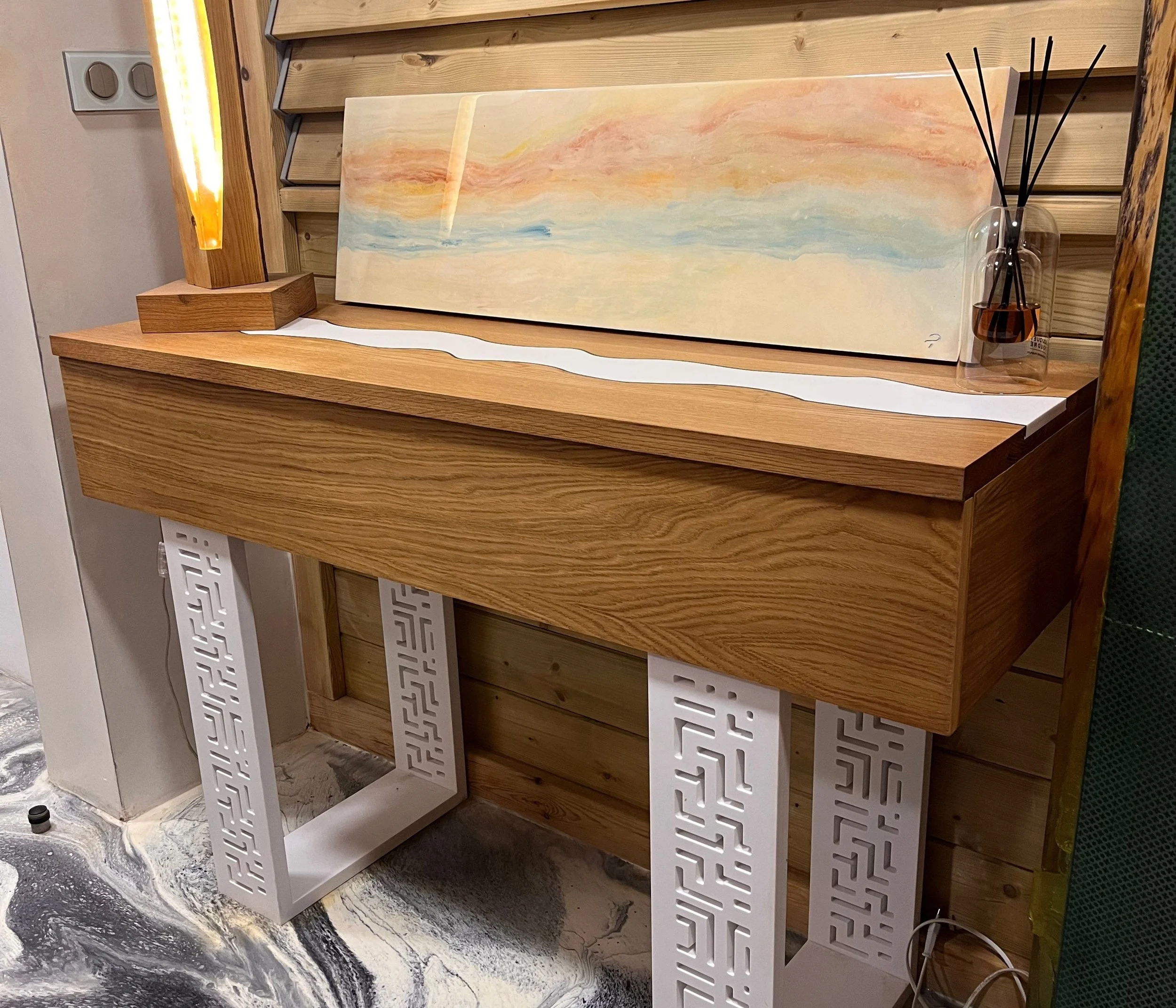 Table en bois avec une sculpture en bois et une peinture abstraite colorée en arrière-plan, un diffuseur d'huiles essentielles avec des bâtonnets, sur une surface en marbre gris et blanc.