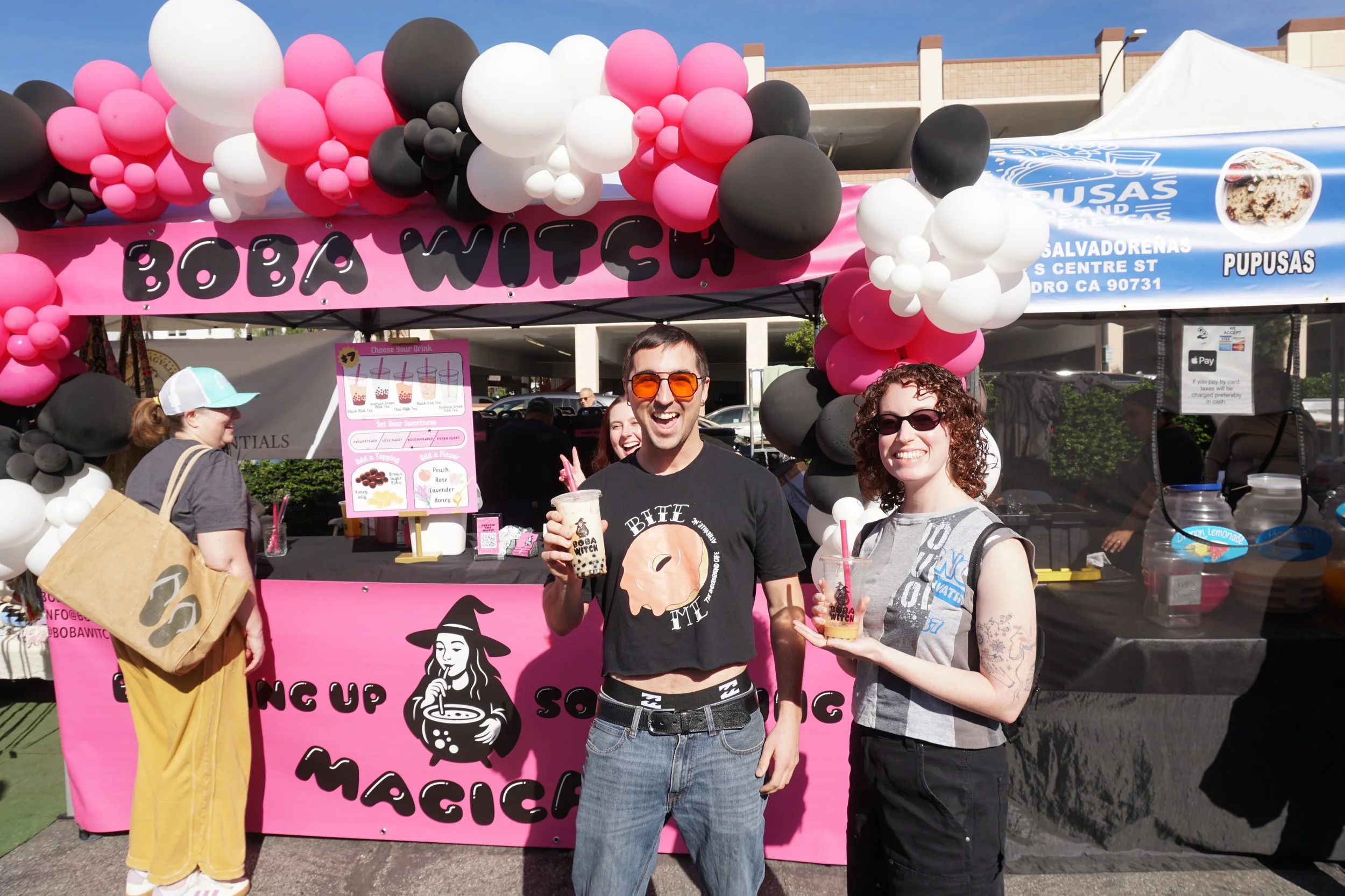 Two people smiling and holding beverages at a colorful outdoor booth decorated with pink, white, and black balloons, with a pink banner and a menu for bubble tea.