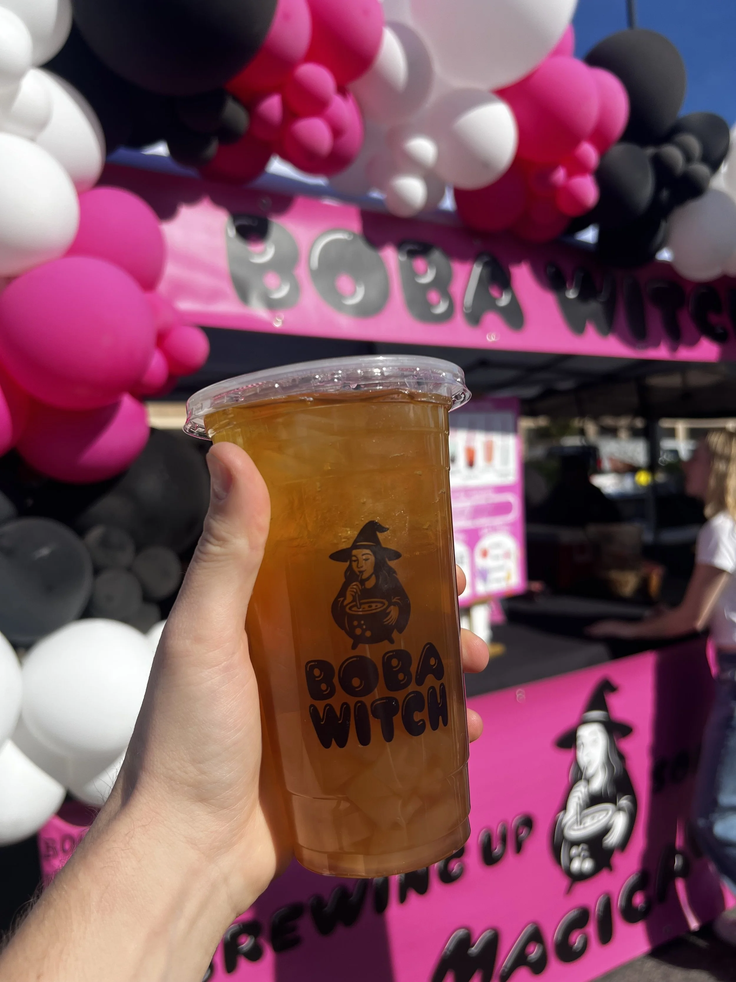 Hand holding a large iced drink in a clear cup with a witch logo and the words 'Boba Witch.' In the background, there is a pink and black balloon arch and a pink sign for a Boba Tea stand.