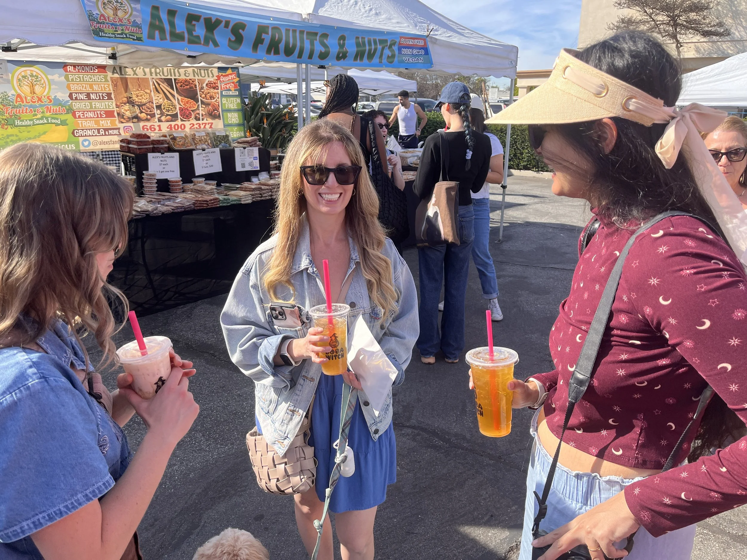 Three women standing outdoors at a market, drinking colorful beverages with pink straws, while chatting and smiling. In the background, there are market stalls and other people.