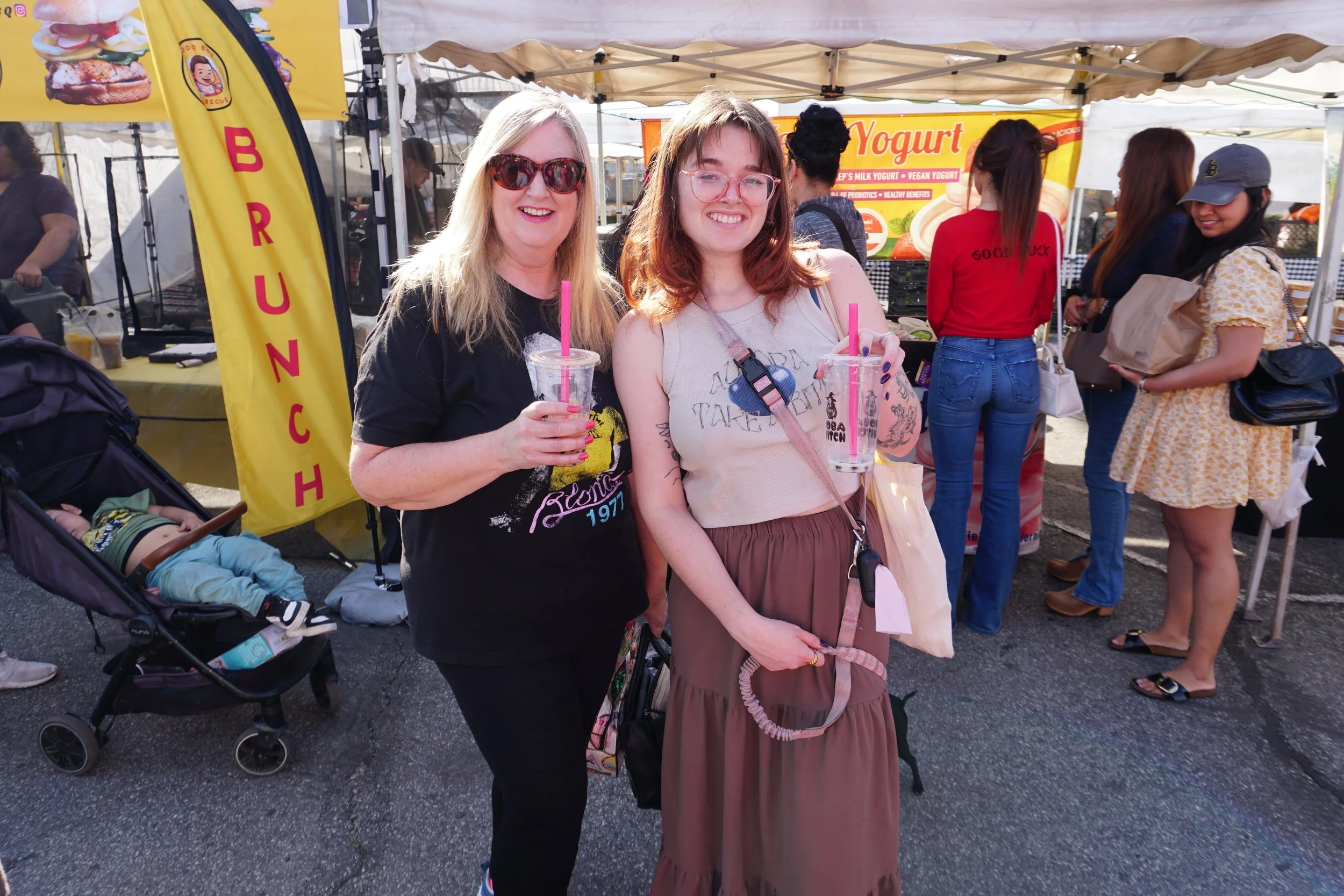 Two women smiling and holding drinks at an outdoor market or festival with a yellow banner and tents in the background. One woman has blonde hair and sunglasses, while the other has red hair and glasses.